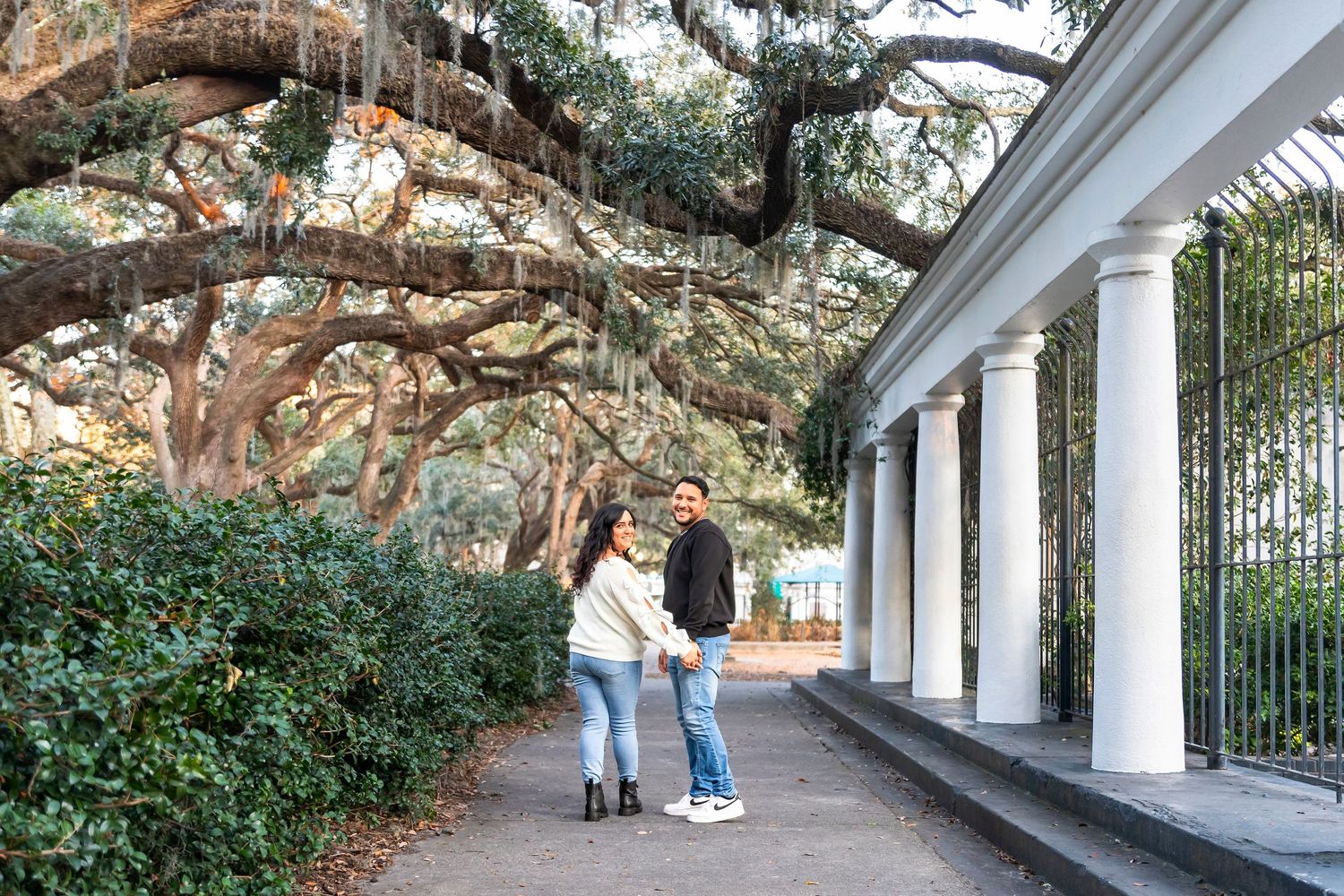 People walking along a covered walkway with white columns and Spanish moss hanging from large oak trees overhead.