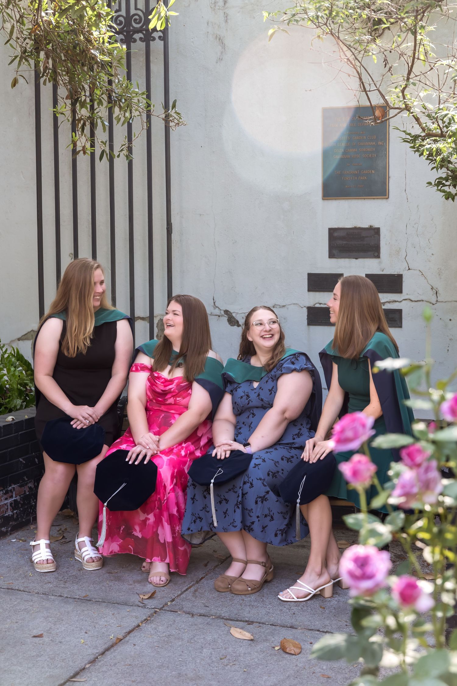 Four female graduates in green academic hoods laugh together on a bench surrounded by pink flowers.