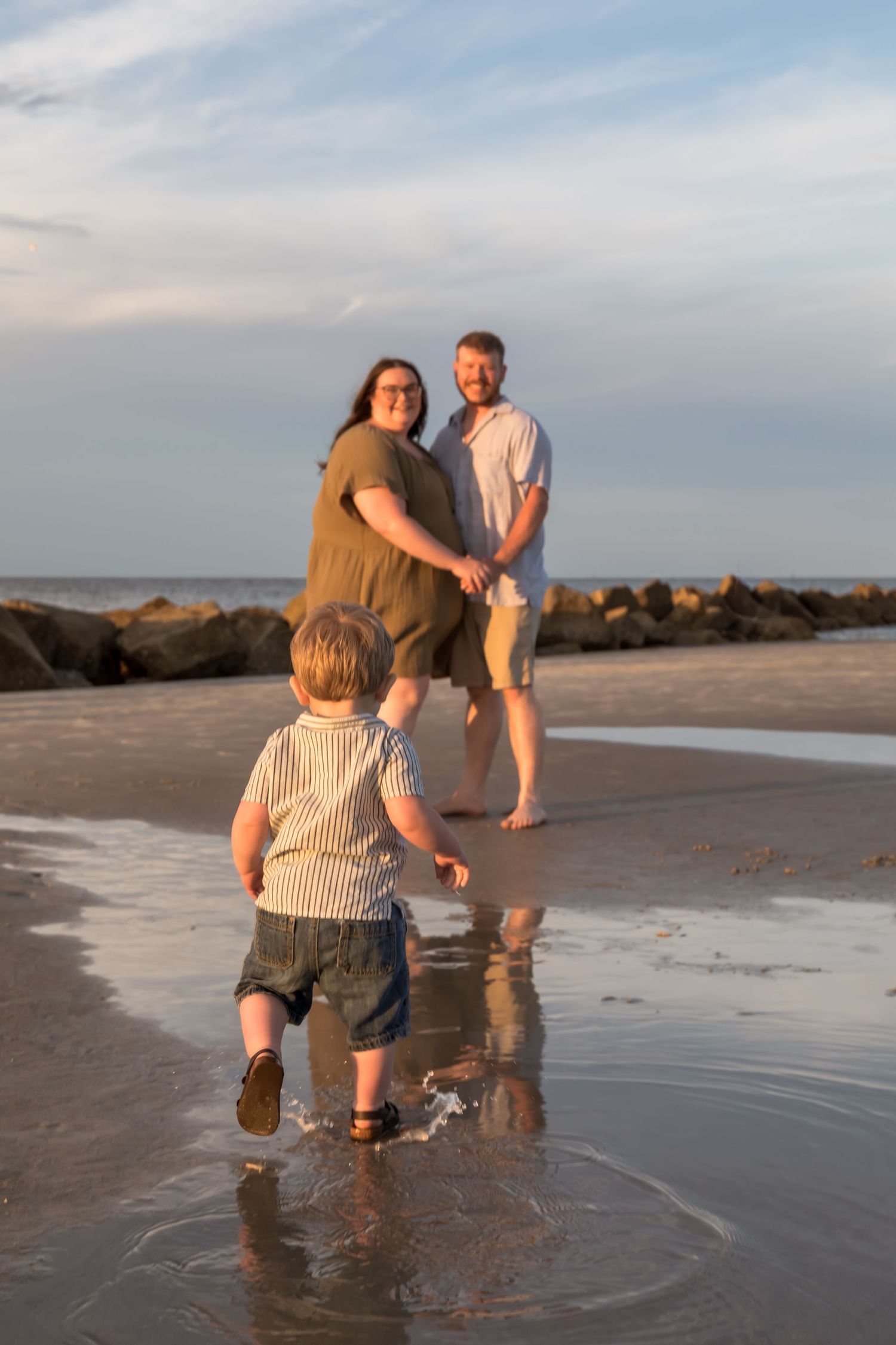 A young family enjoys a golden hour beach walk, with a toddler splashing toward his smiling parents near rocky shores.