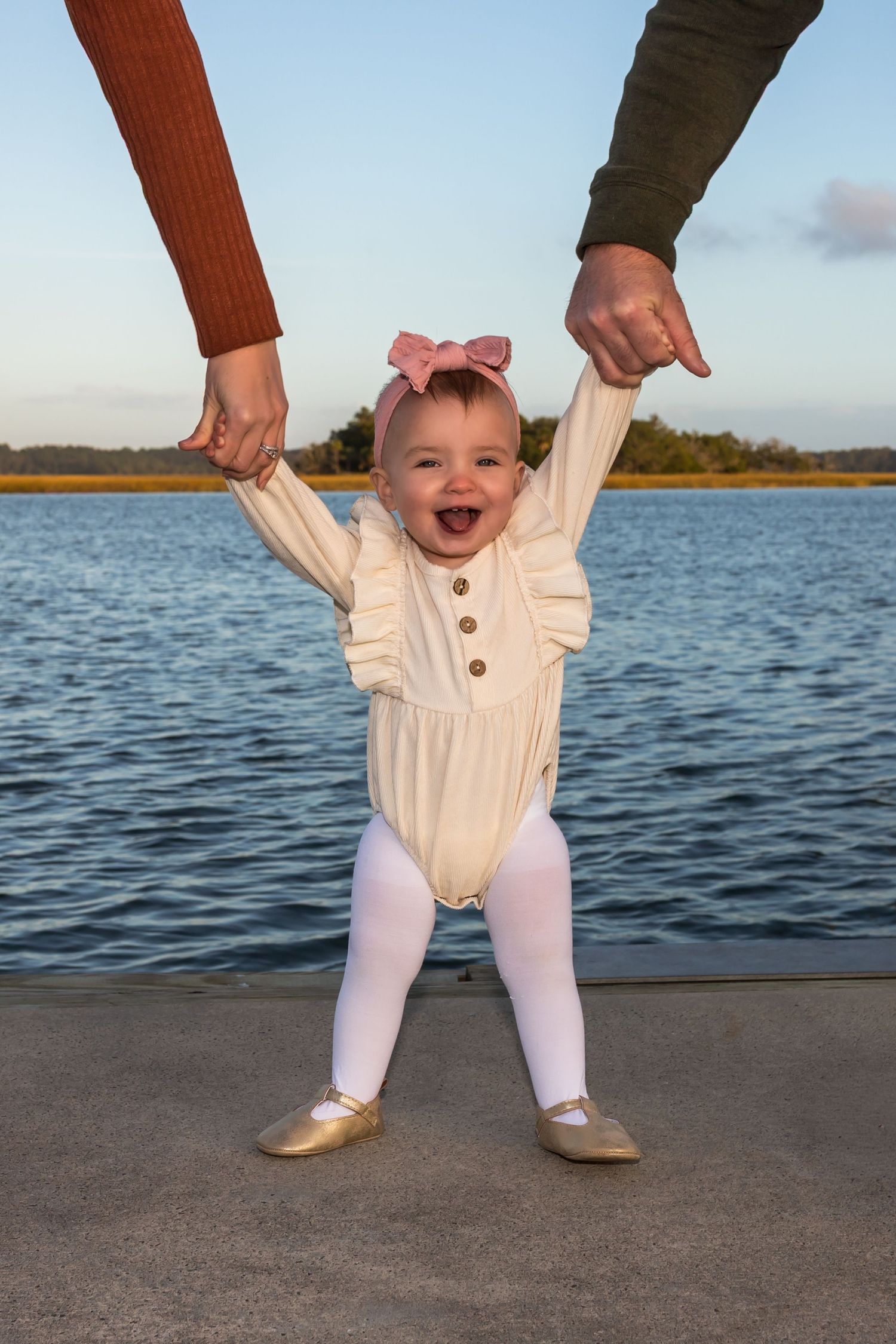 Baby in white outfit learning to walk on waterfront dock.