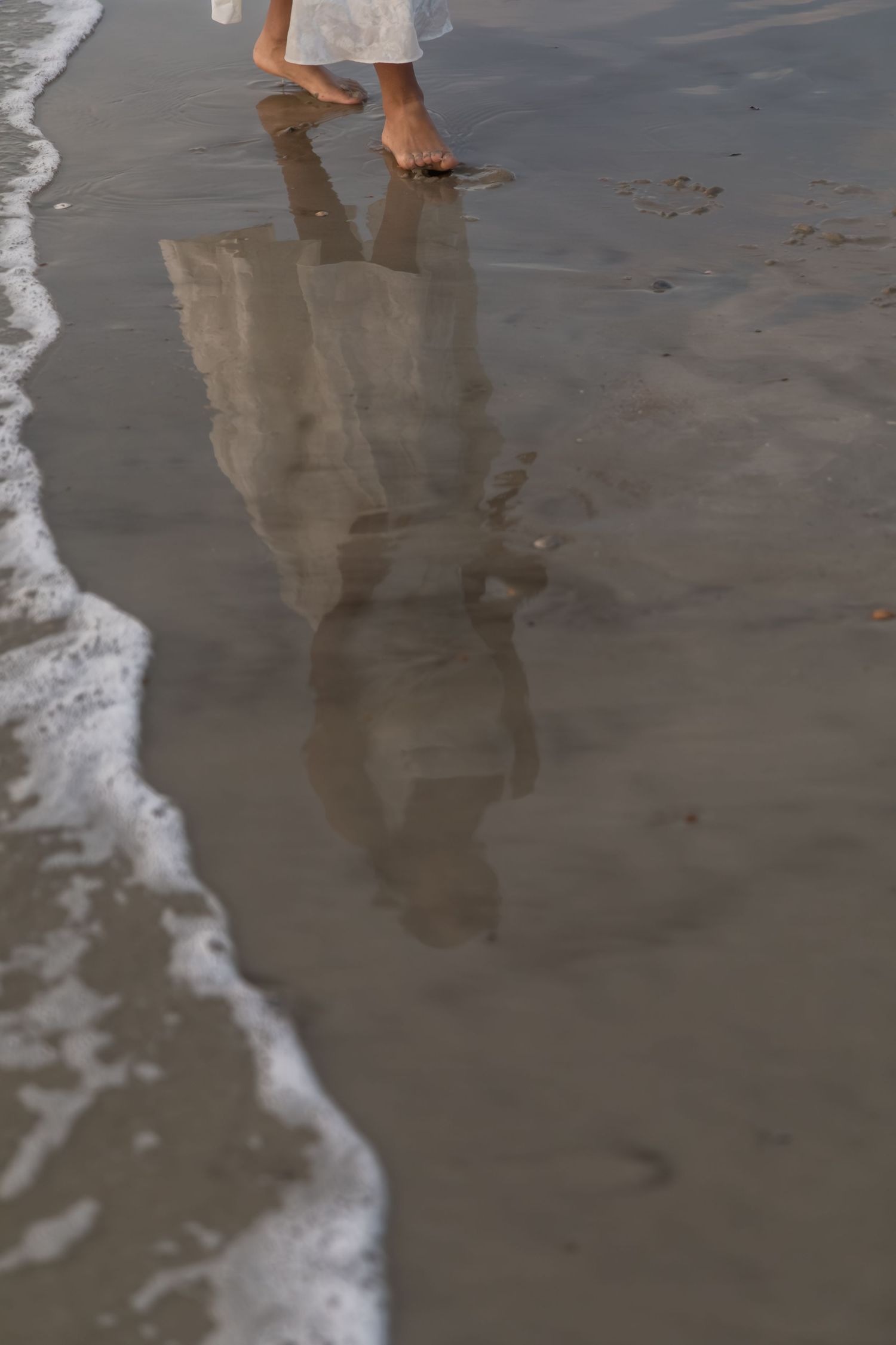 Barefoot person in white dress walking on wet sandy beach with reflection visible in shallow water near ocean waves.