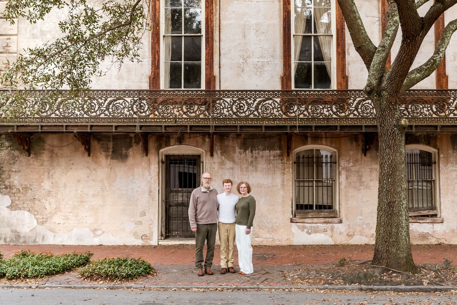 Historic brick building facade with ornate wrought iron balconies and tall windows under a leafy tree.