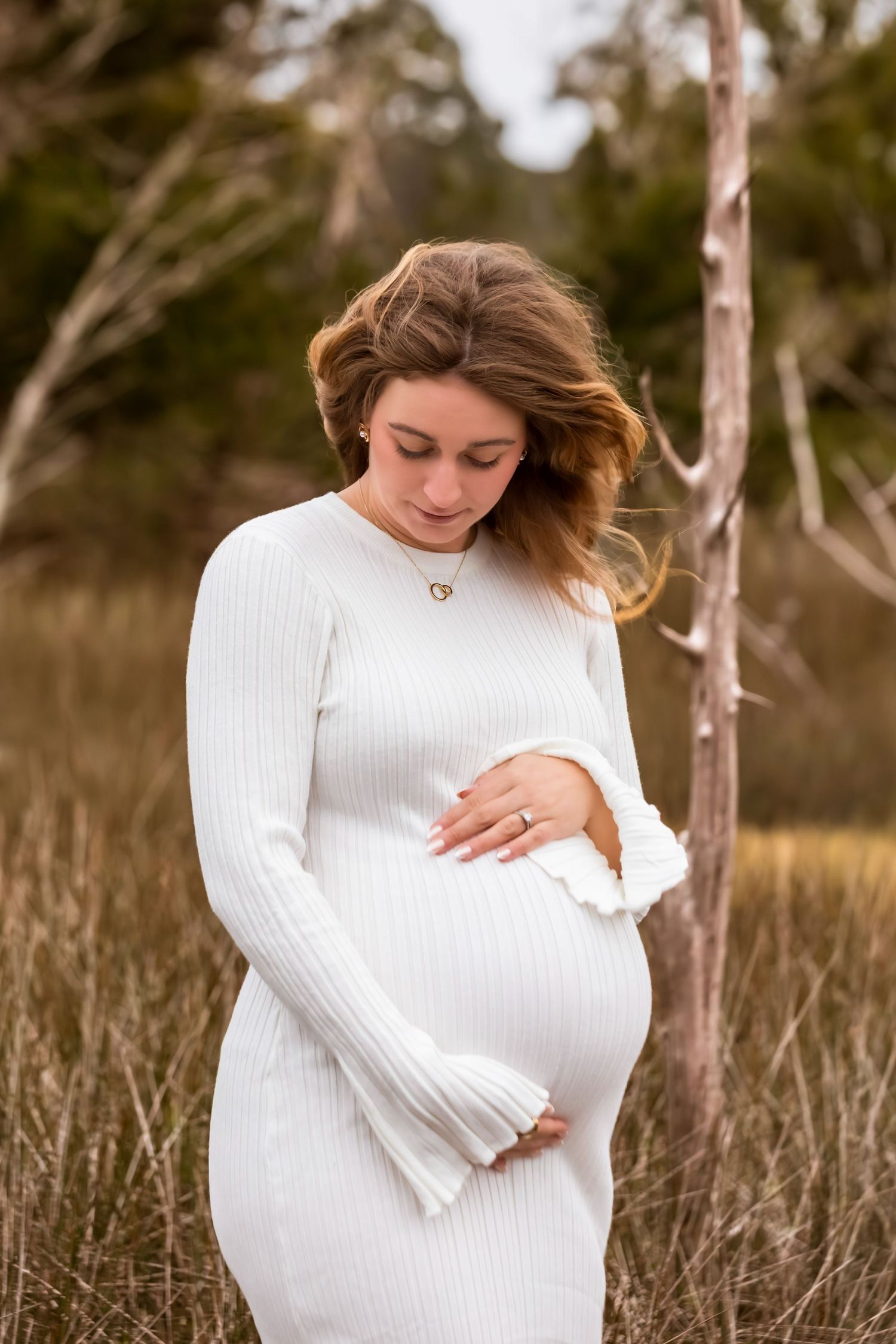 A person in a white sweater poses in a natural field setting during a maternity photoshoot.