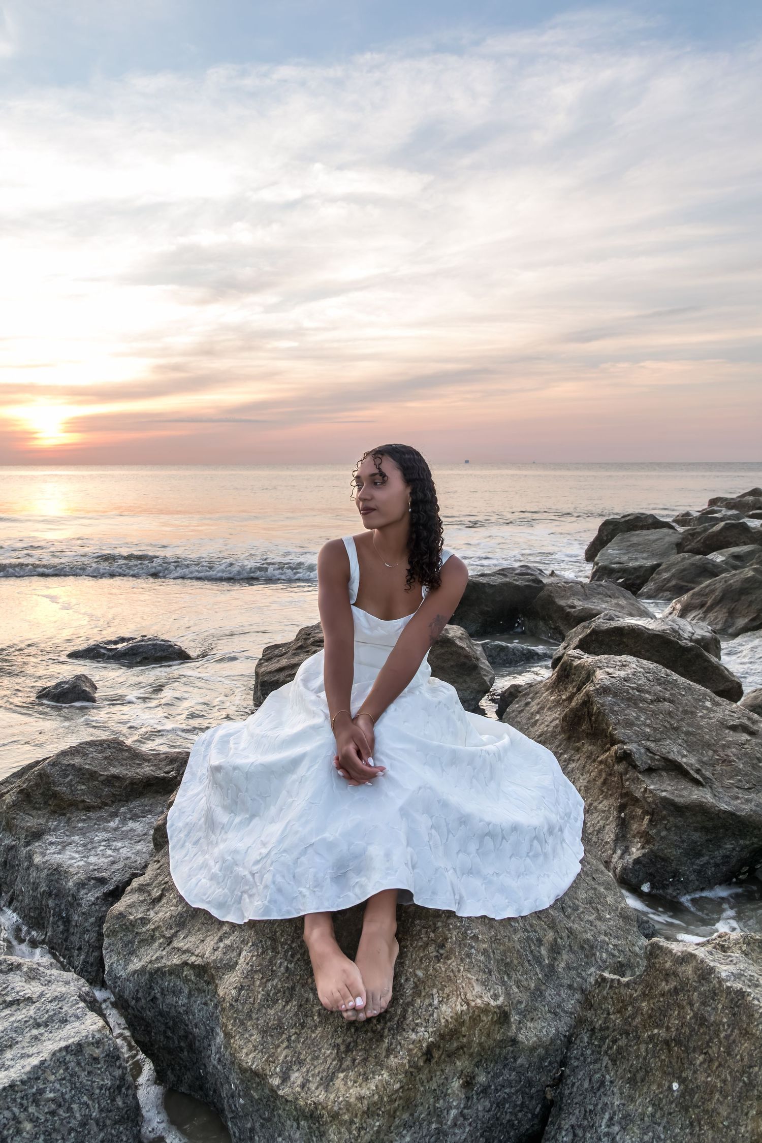 Woman in white sundress sits on rocky shore gazing at colorful ocean sunset.