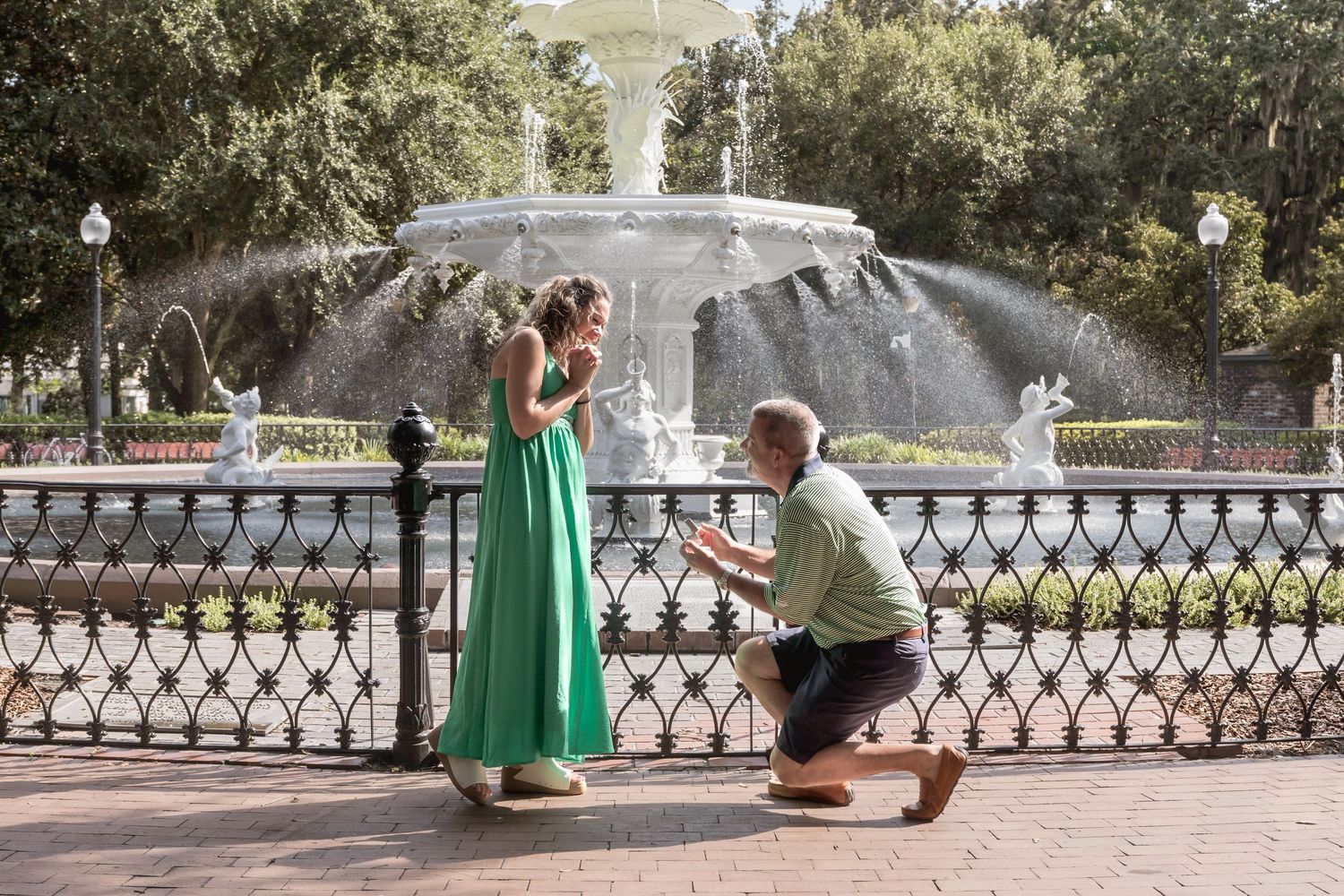 Man proposing to woman in green dress by ornate fountain in sunny park setting.