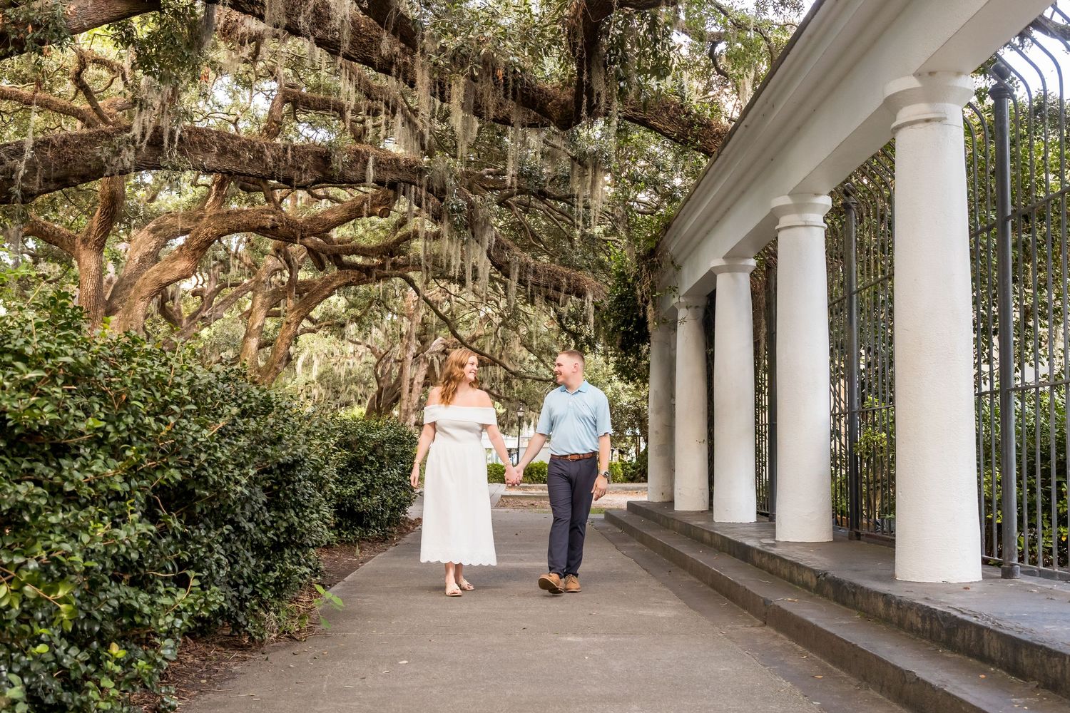 A couple poses together under a white columned pergola in a romantic garden setting.