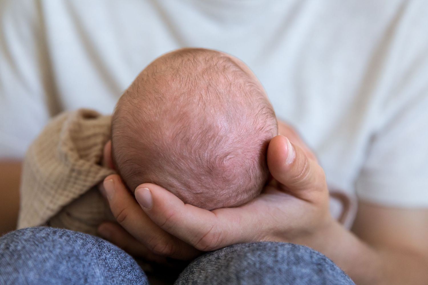 Parent cradling newborn baby's bald head in both hands, close-up tender moment of bonding.