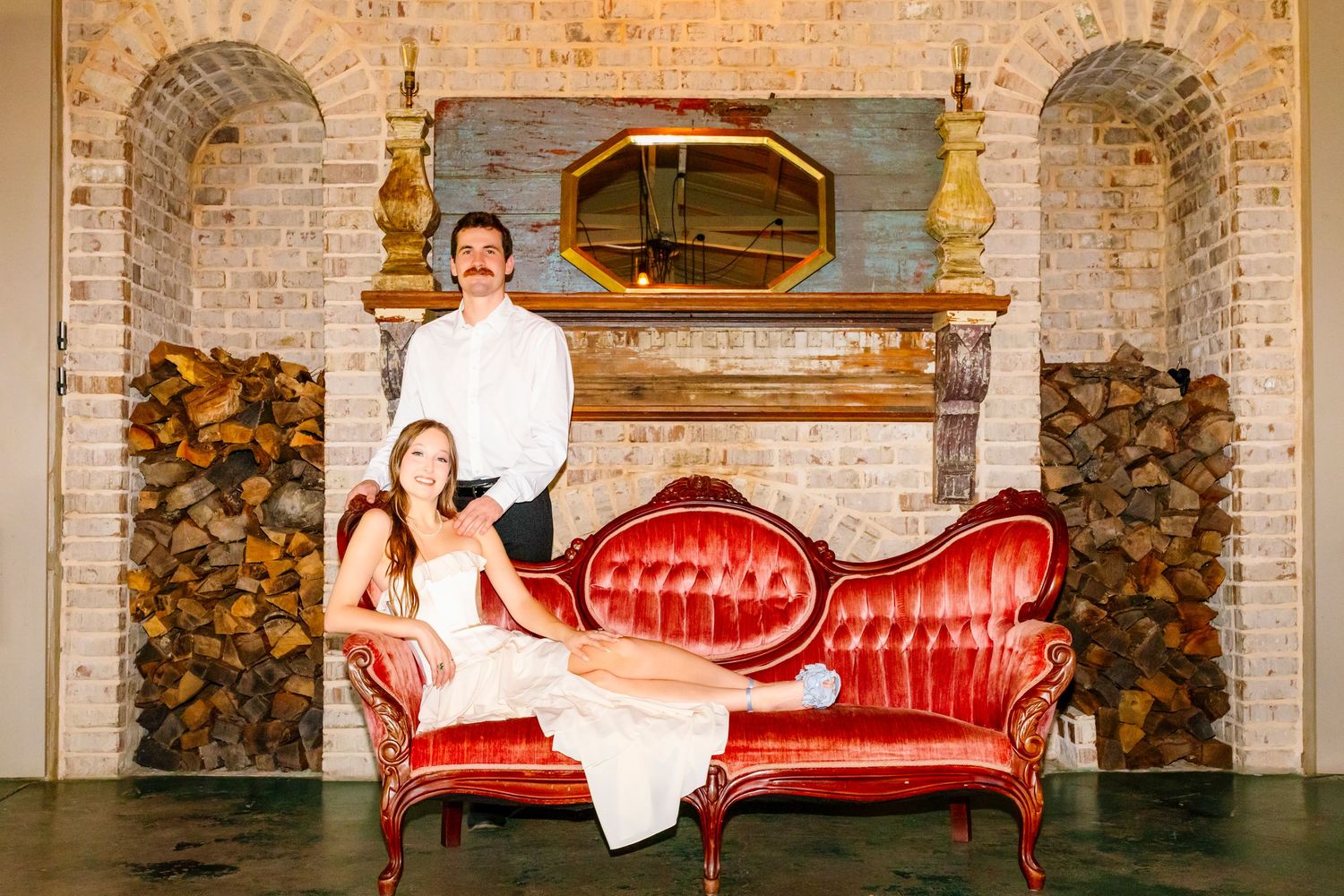 Elegant indoor photo session with vintage red chaise lounge against brick wall.