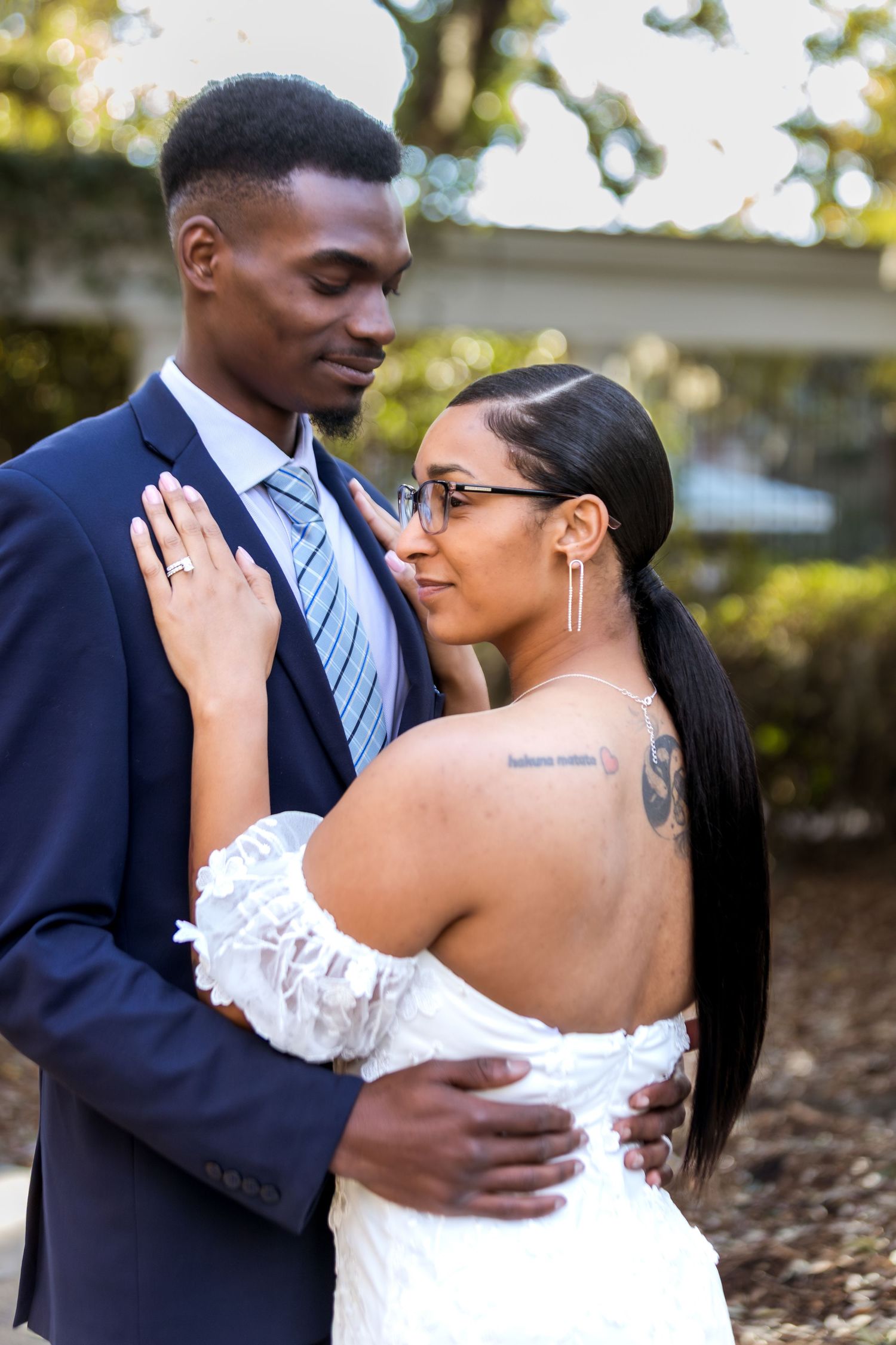 A couple in wedding attire embrace outdoors, the bride in a white off-shoulder gown and groom in a navy suit.