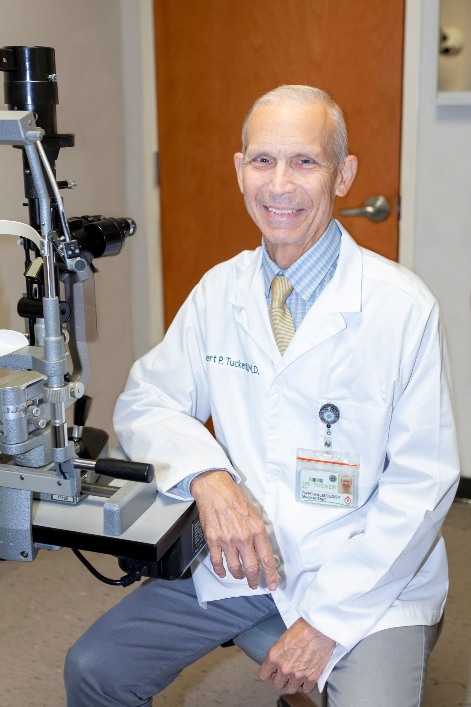 Smiling male doctor in white lab coat seated next to slit lamp equipment in medical examination room.