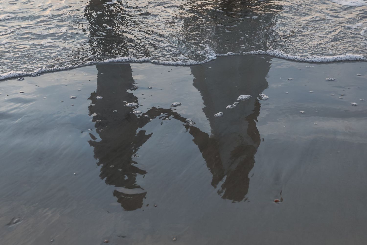 Reflections of two people standing on a wet sandy beach shore with gentle ocean waves.
