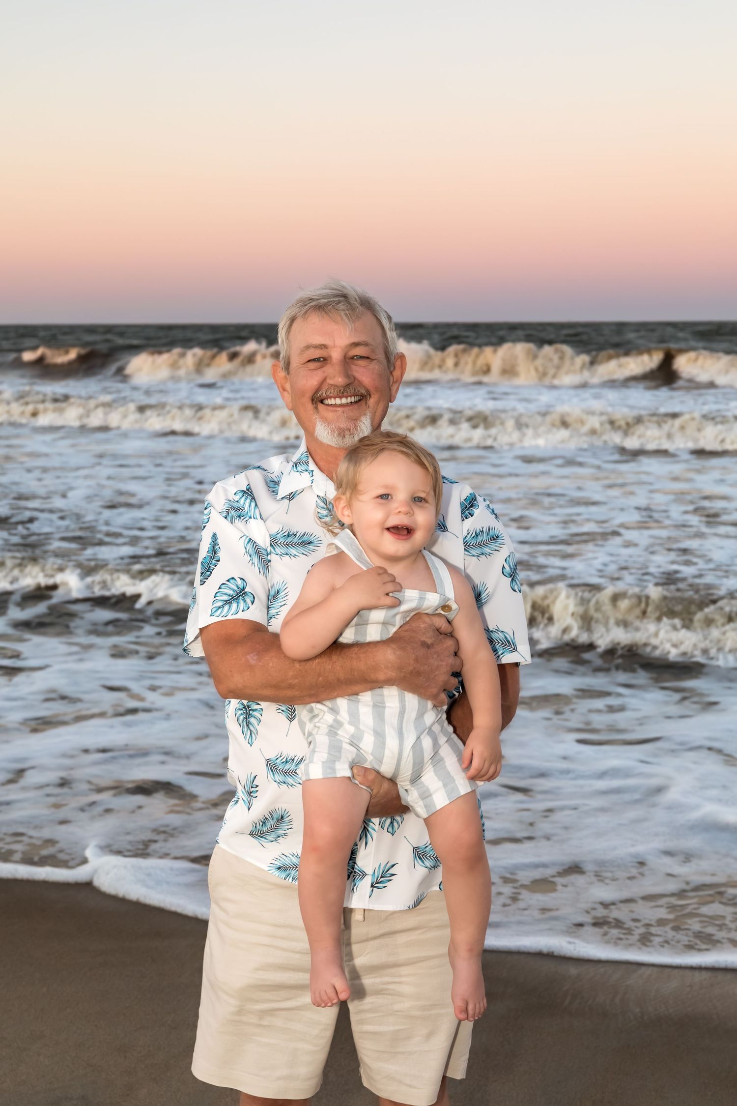 Smiling grandfather holding happy baby on beach at sunset, waves crashing behind them.