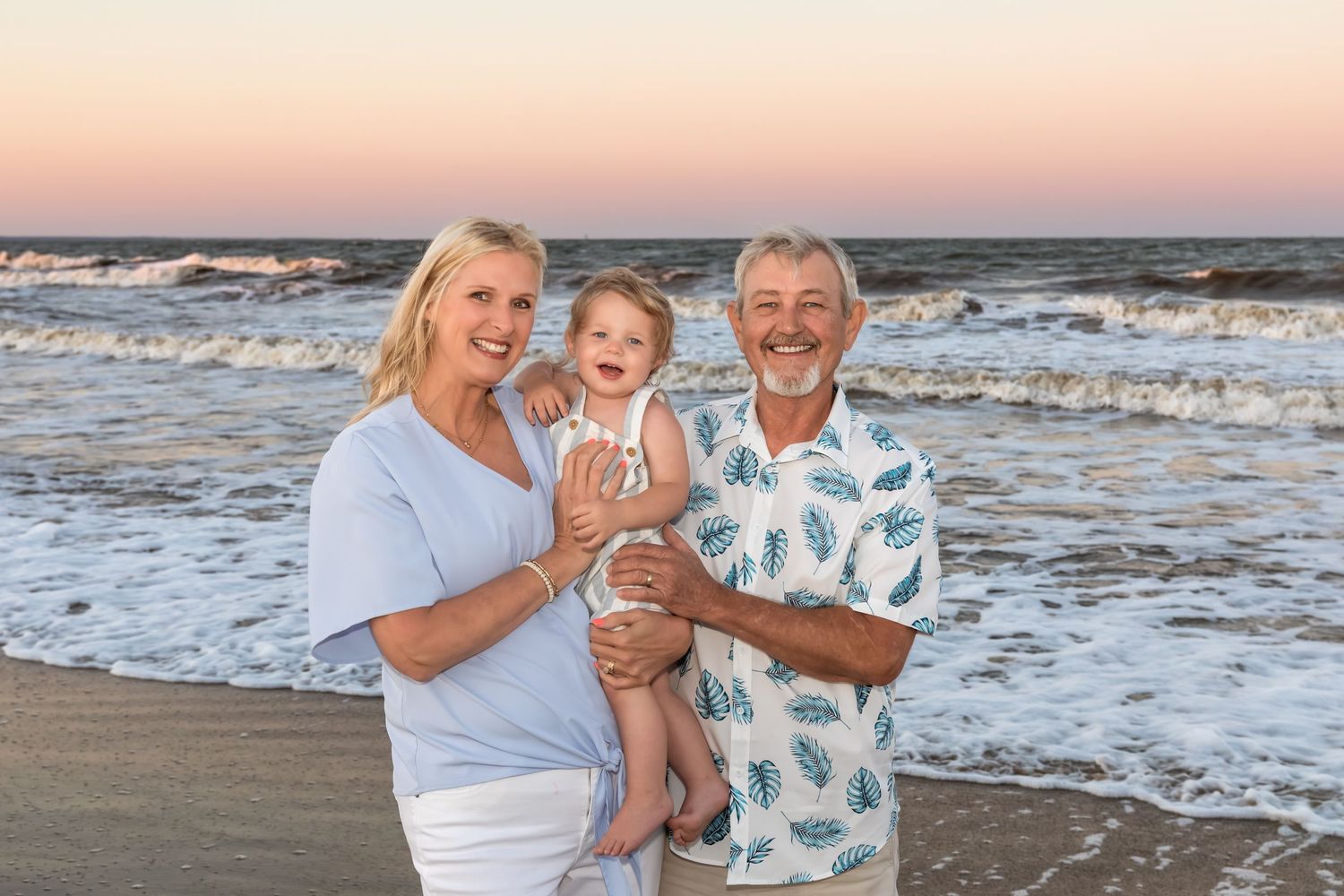 Grandparents smiling and holding a baby on a beach at sunset with ocean waves in the background.