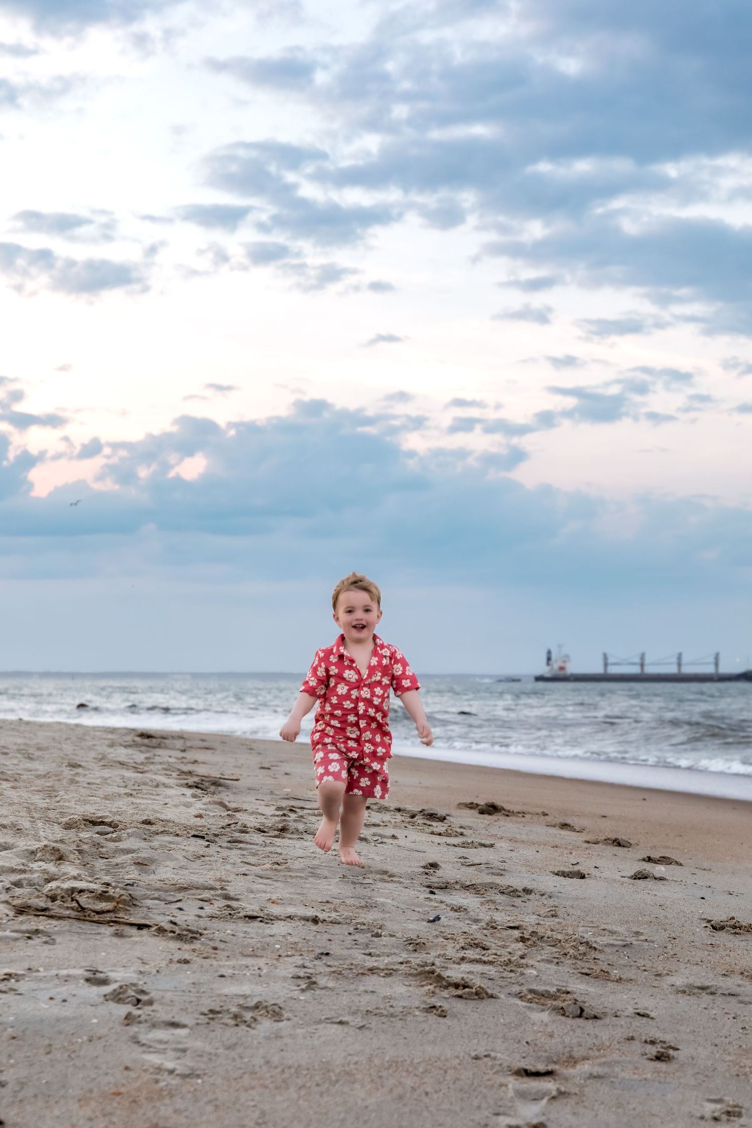 A joyful toddler in a red floral outfit runs barefoot along a sandy beach with ocean waves and cloudy sky behind.