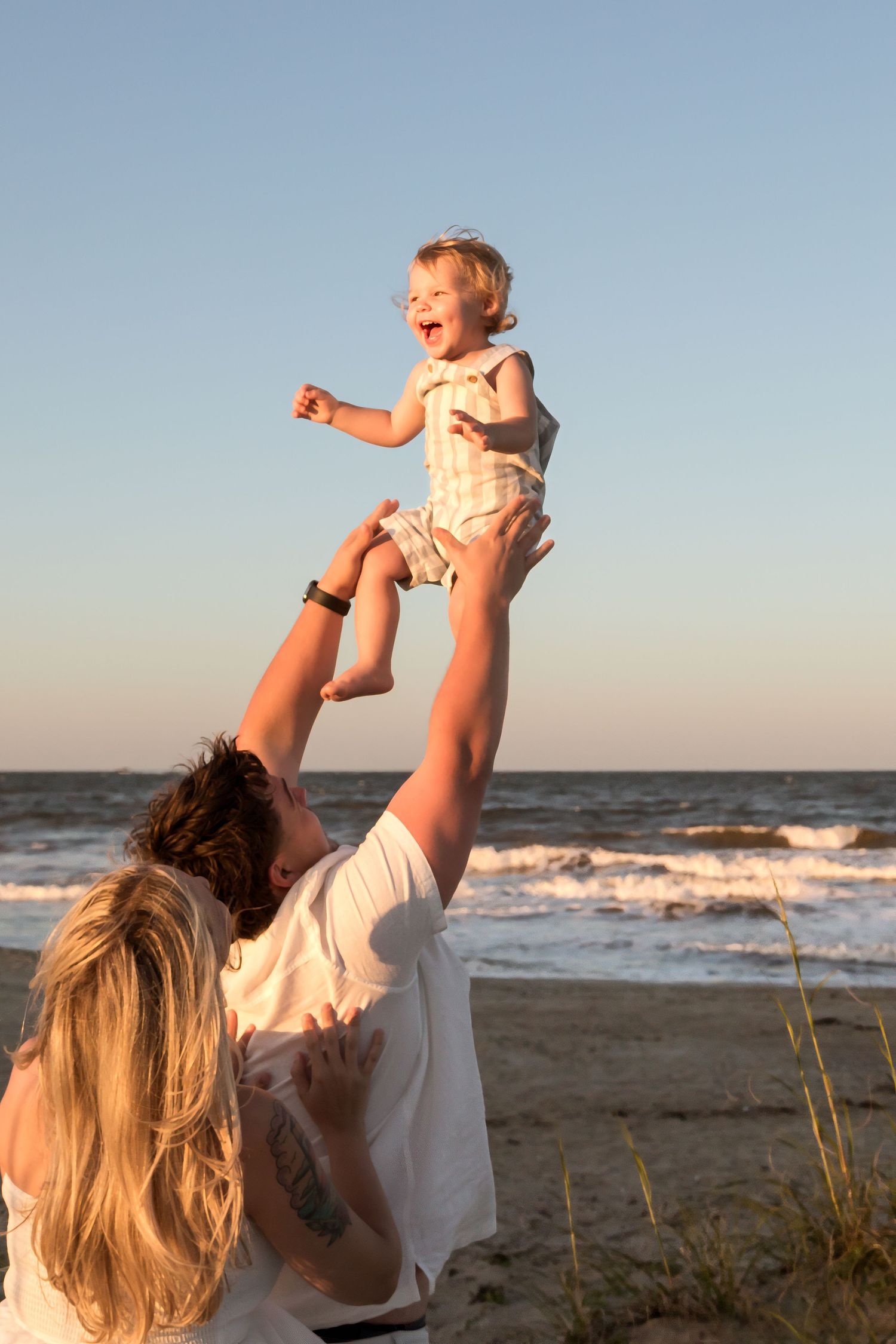 Parent lifting laughing toddler high in the air on a sunny beach at golden hour.