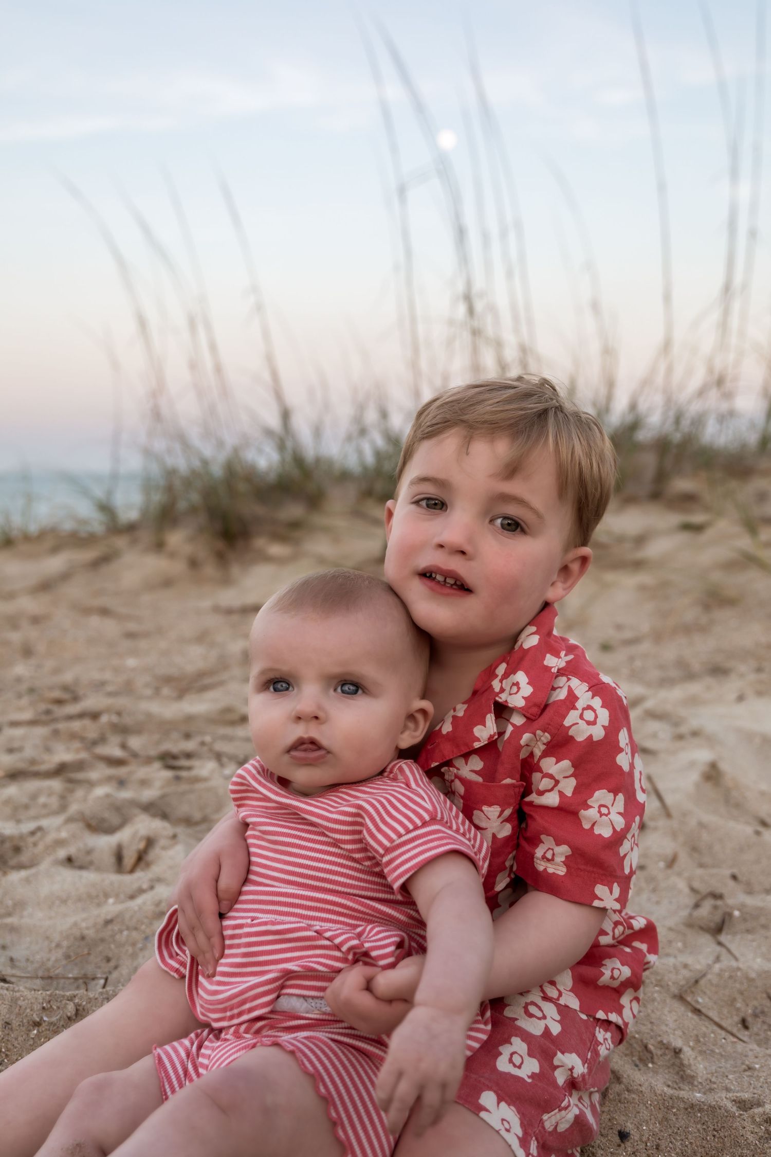 Two young siblings sit together on sandy beach, older boy in floral shirt hugging baby in pink stripes at dusk.