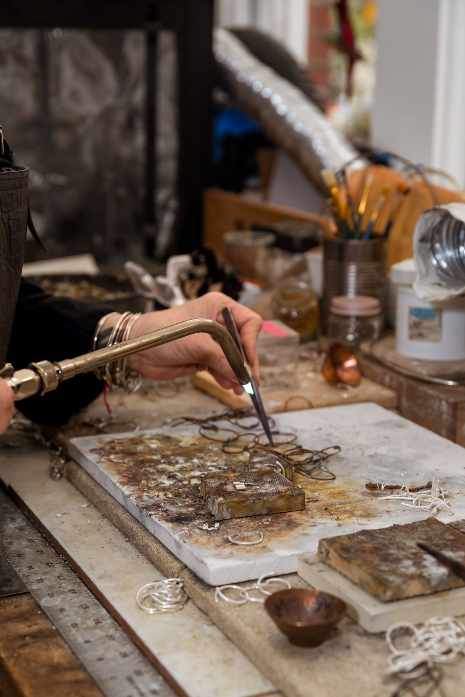 Jeweler using a torch to solder metal rings on a workbench cluttered with tools and materials in a craft studio.