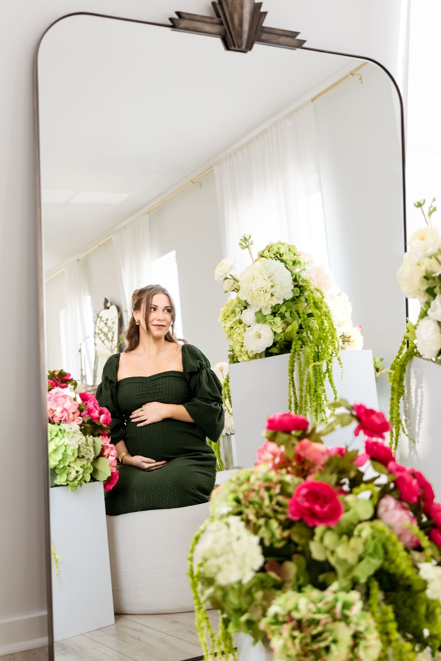Pregnant woman in green dress reflected in mirror surrounded by lush floral arrangements in white vases.