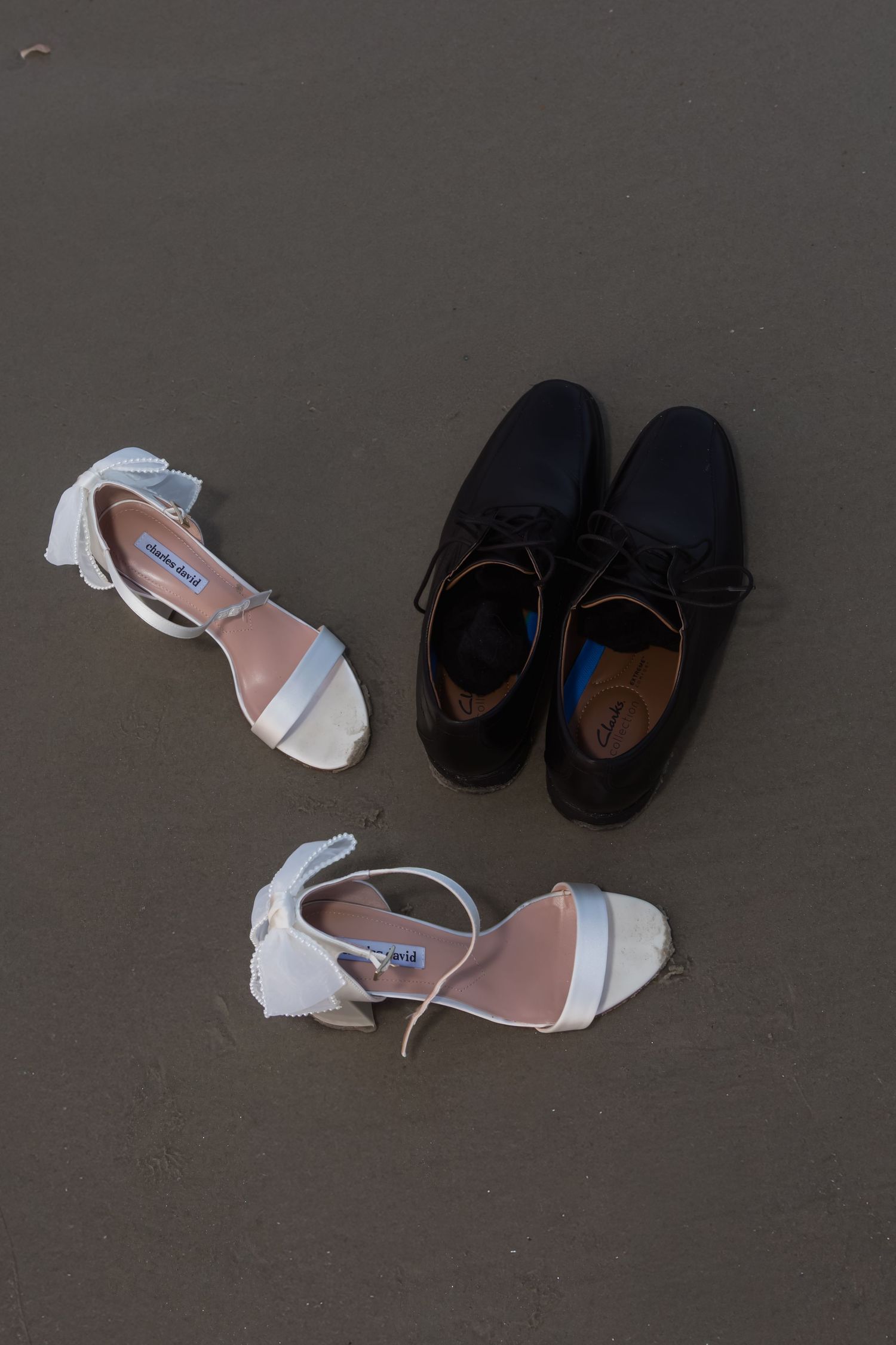 White bridal heels and black groom dress shoes placed on sandy beach for wedding day flat lay photo.