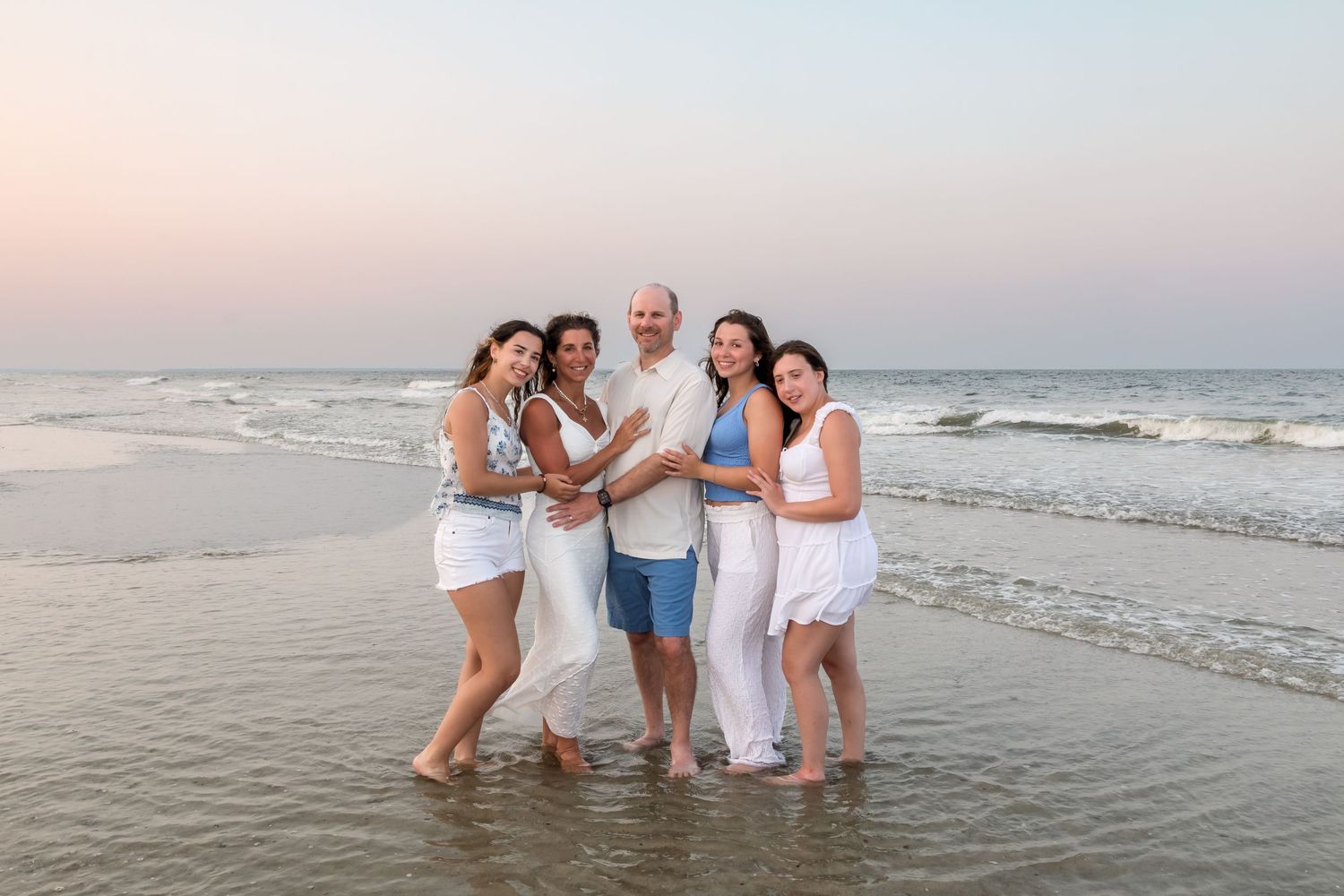 Family of five posing together on a sandy beach at sunset, wearing white and blue outfits with ocean waves behind them.