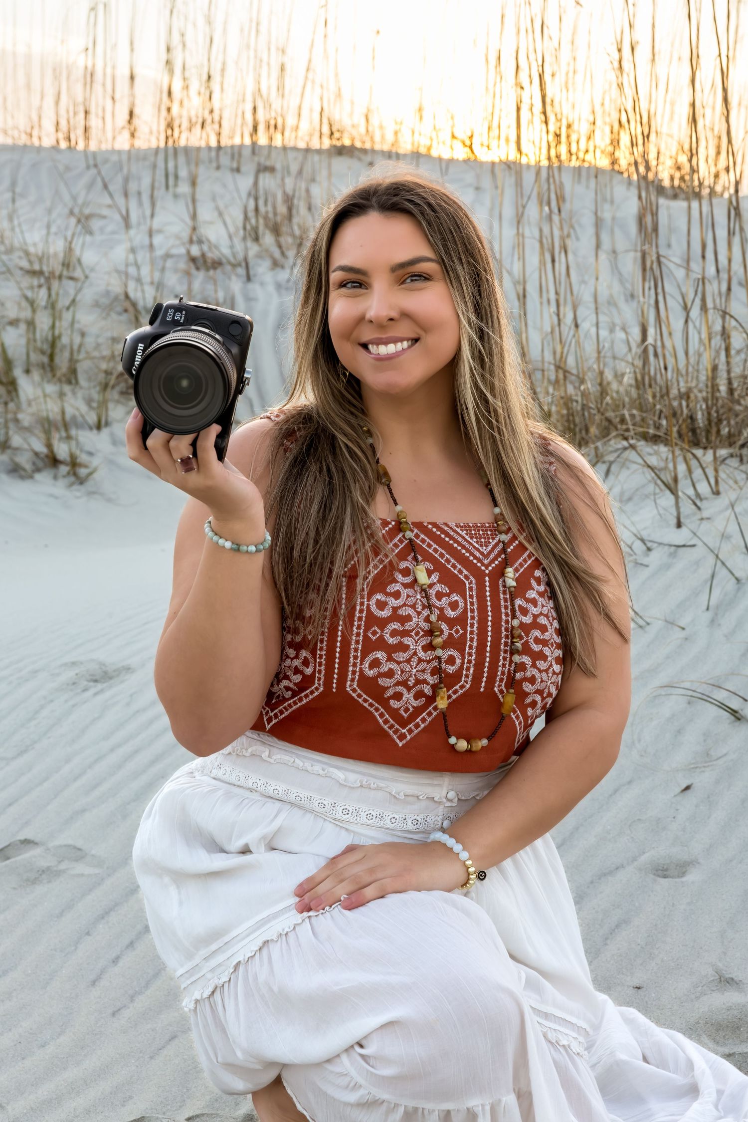 A photographer in a red embroidered top sits on white sand dunes during sunset while holding a camera.