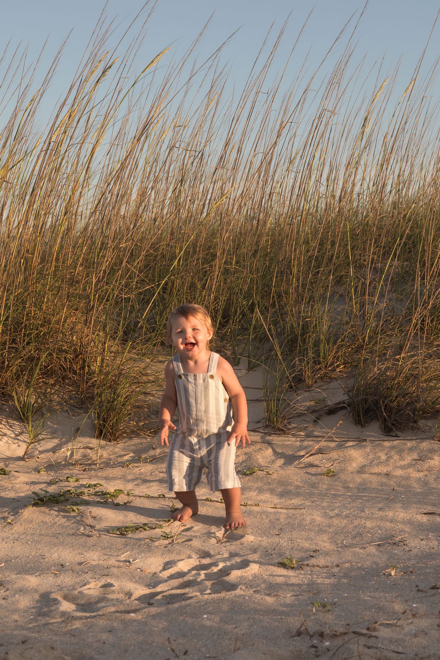 Happy toddler laughing and standing barefoot on beach sand near tall sea grass.