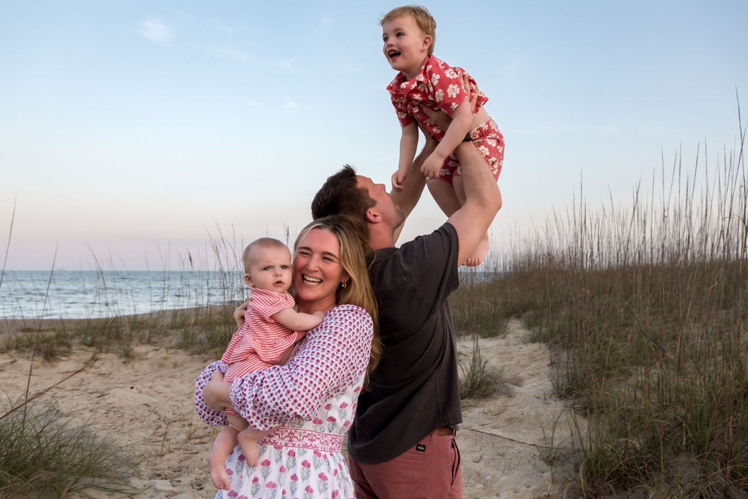 Family of four enjoying a beach sunset, parents lifting laughing toddler while holding baby near sandy dunes.