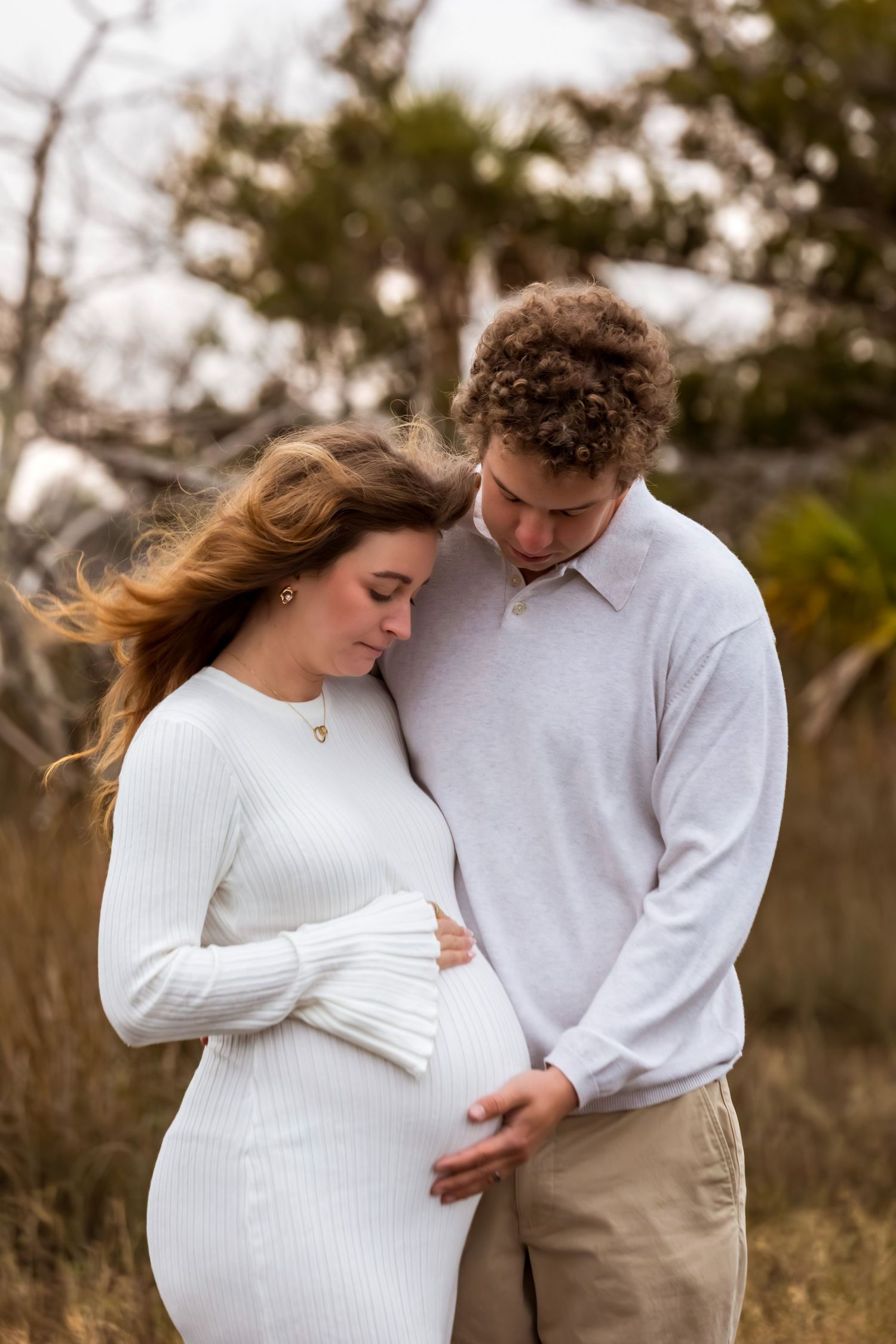 Intimate maternity photo series showing couple in white sweater and khakis embracing in outdoor natural setting.