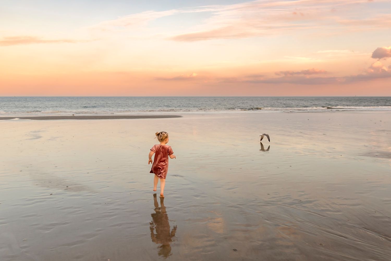 A silhouette stands on a reflective beach at sunset with calm ocean waters and pink-orange sky in the background.