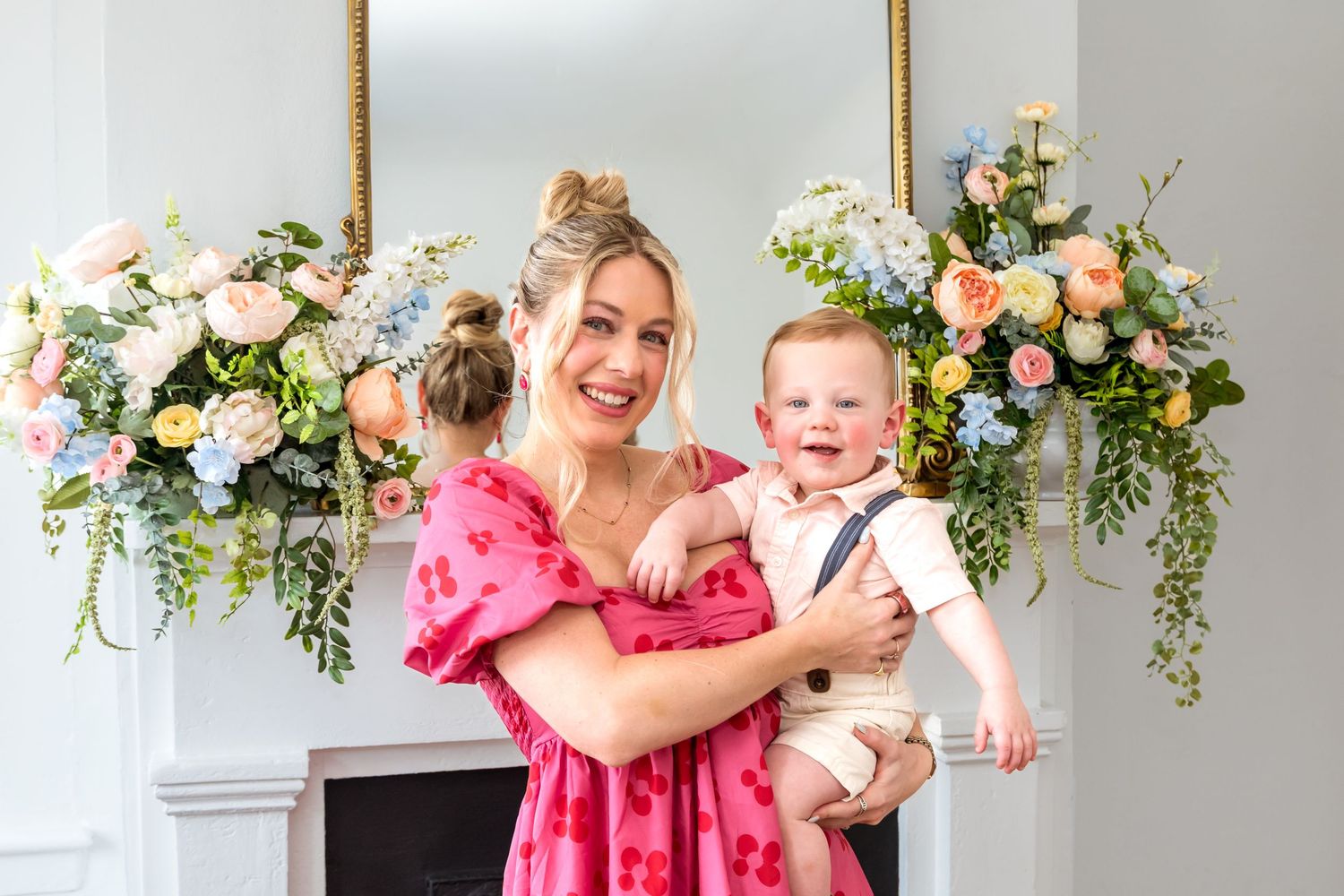 Smiling woman in pink floral dress holding laughing baby boy near floral arrangements on a fireplace mantel.