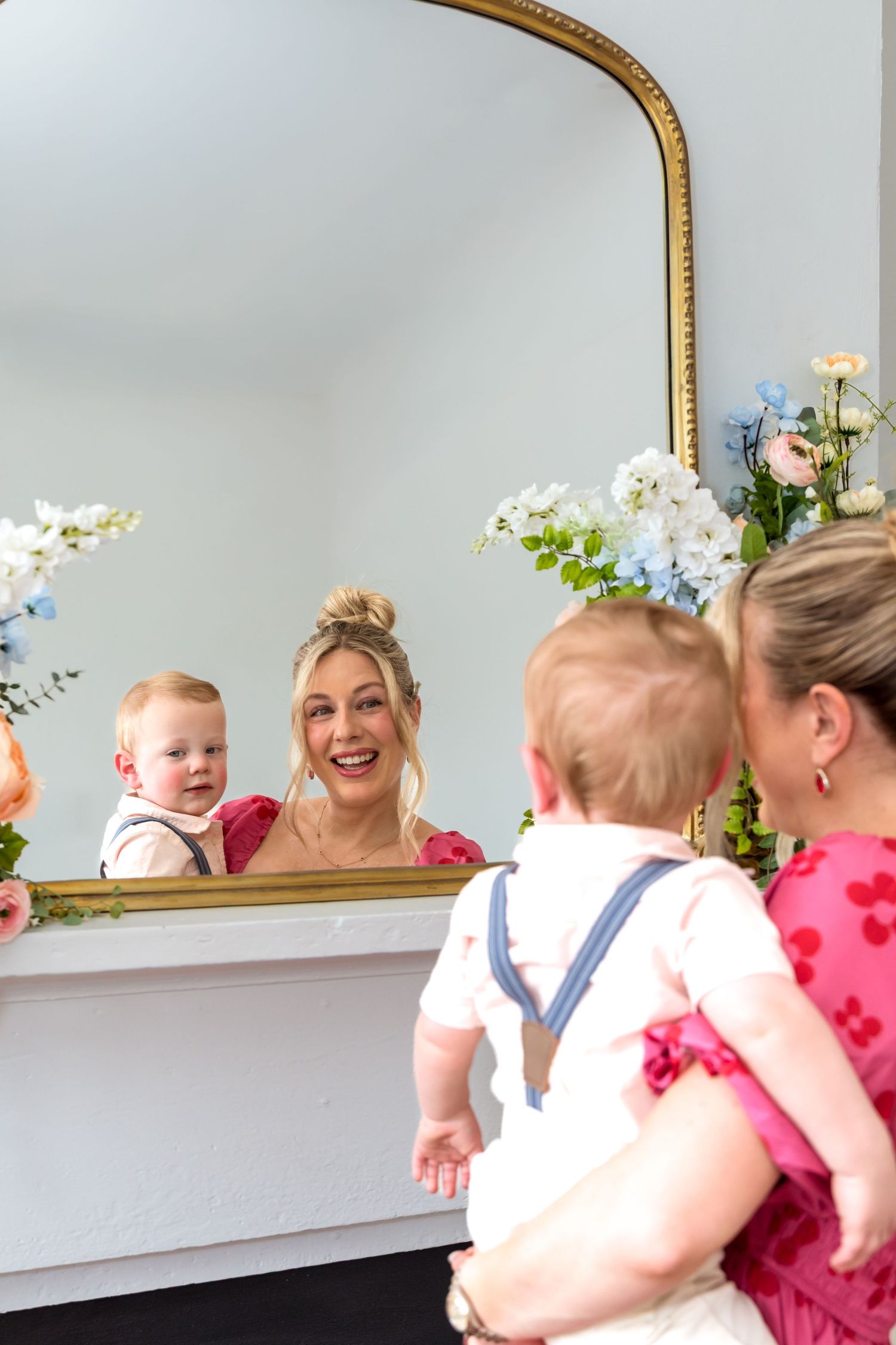 Blonde woman in pink dress holding a baby in suspenders, smiling at their reflection in a gold-framed mirror with flowers.
