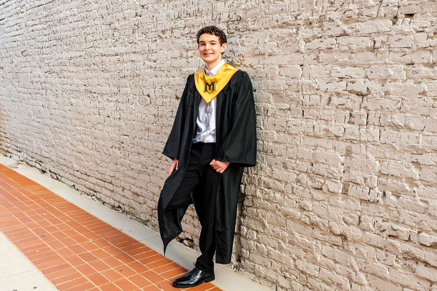 Graduates in black caps and gowns pose against a white brick wall during commencement celebrations.