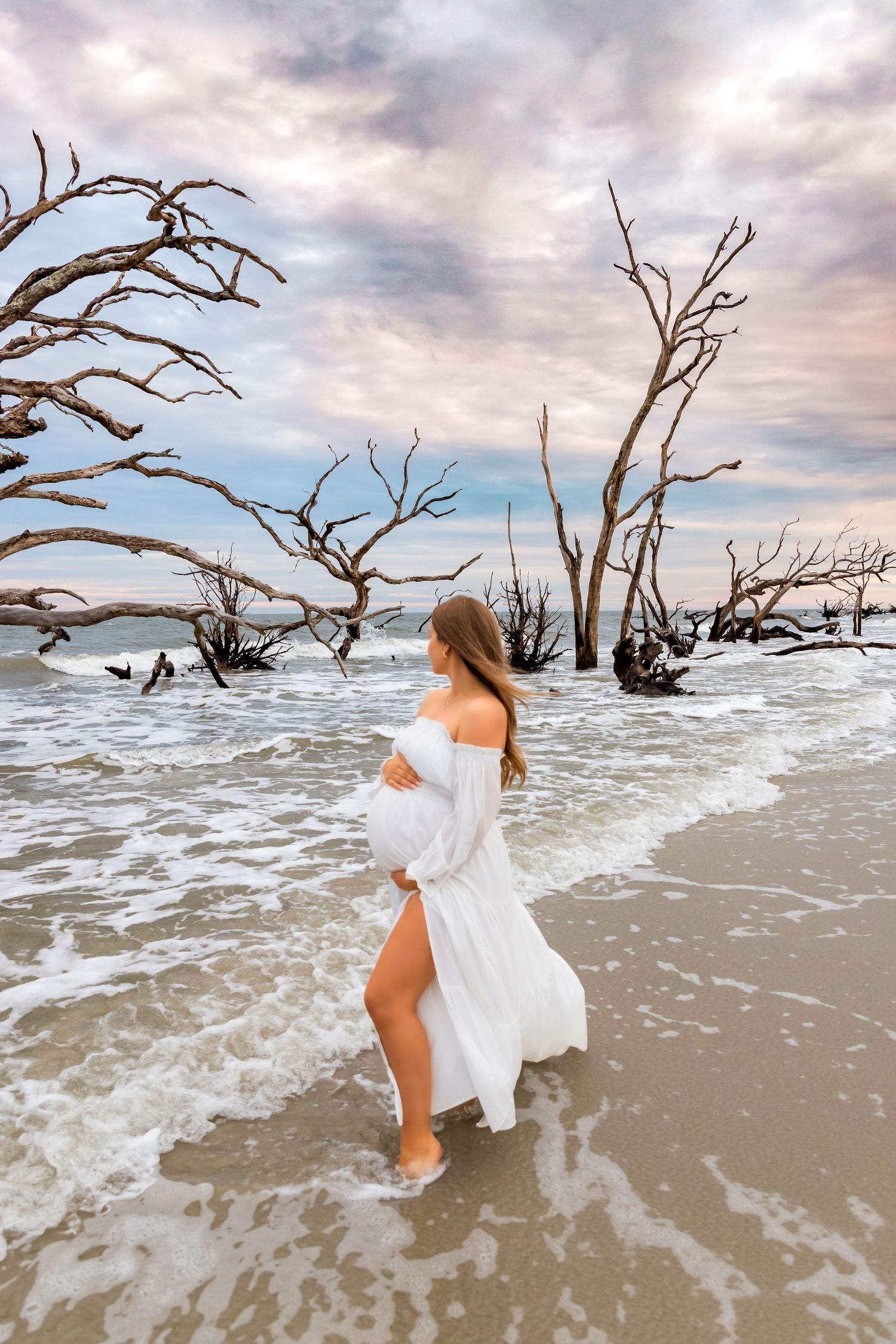 Woman in white dress twirling on beach with driftwood and waves.