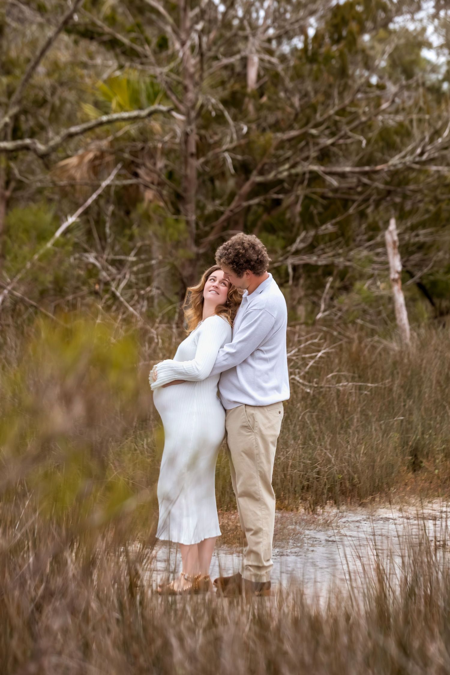 A romantic couple embraces in a white dress and khaki pants among tall grass and bare branches in a natural setting.