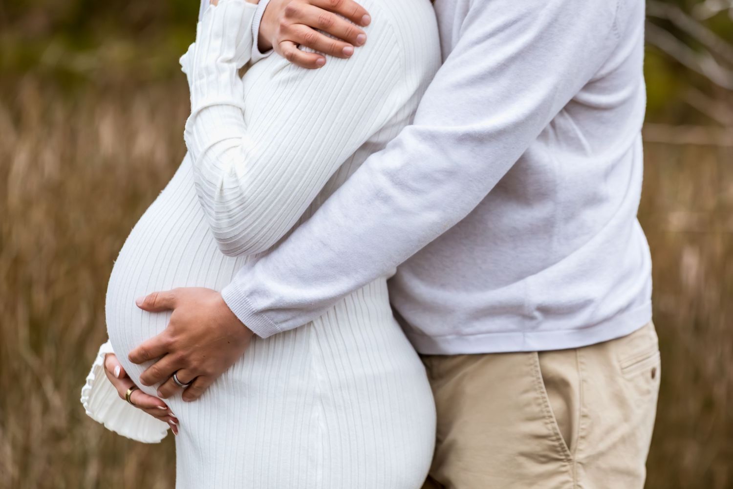 Close up of a couple embracing with hands on a pregnant belly wearing white and khaki clothing.