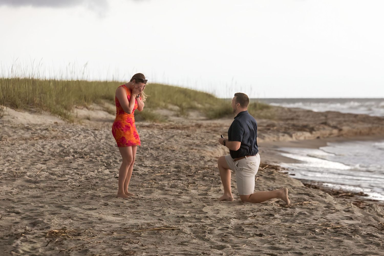 Man kneeling on beach proposing to woman in red dress near ocean shoreline.