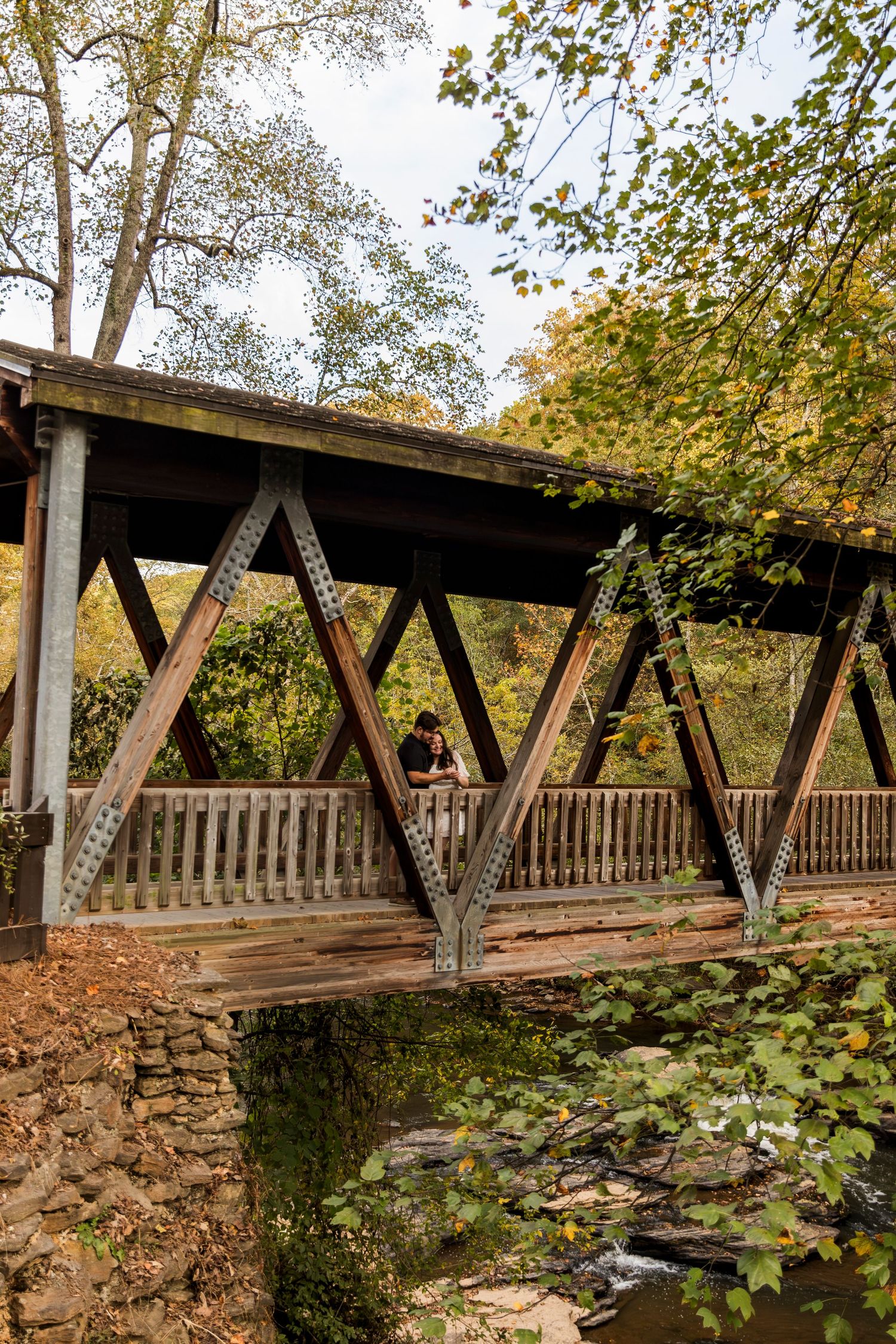 Rustic covered wooden bridge with diagonal support beams surrounded by autumn foliage.
