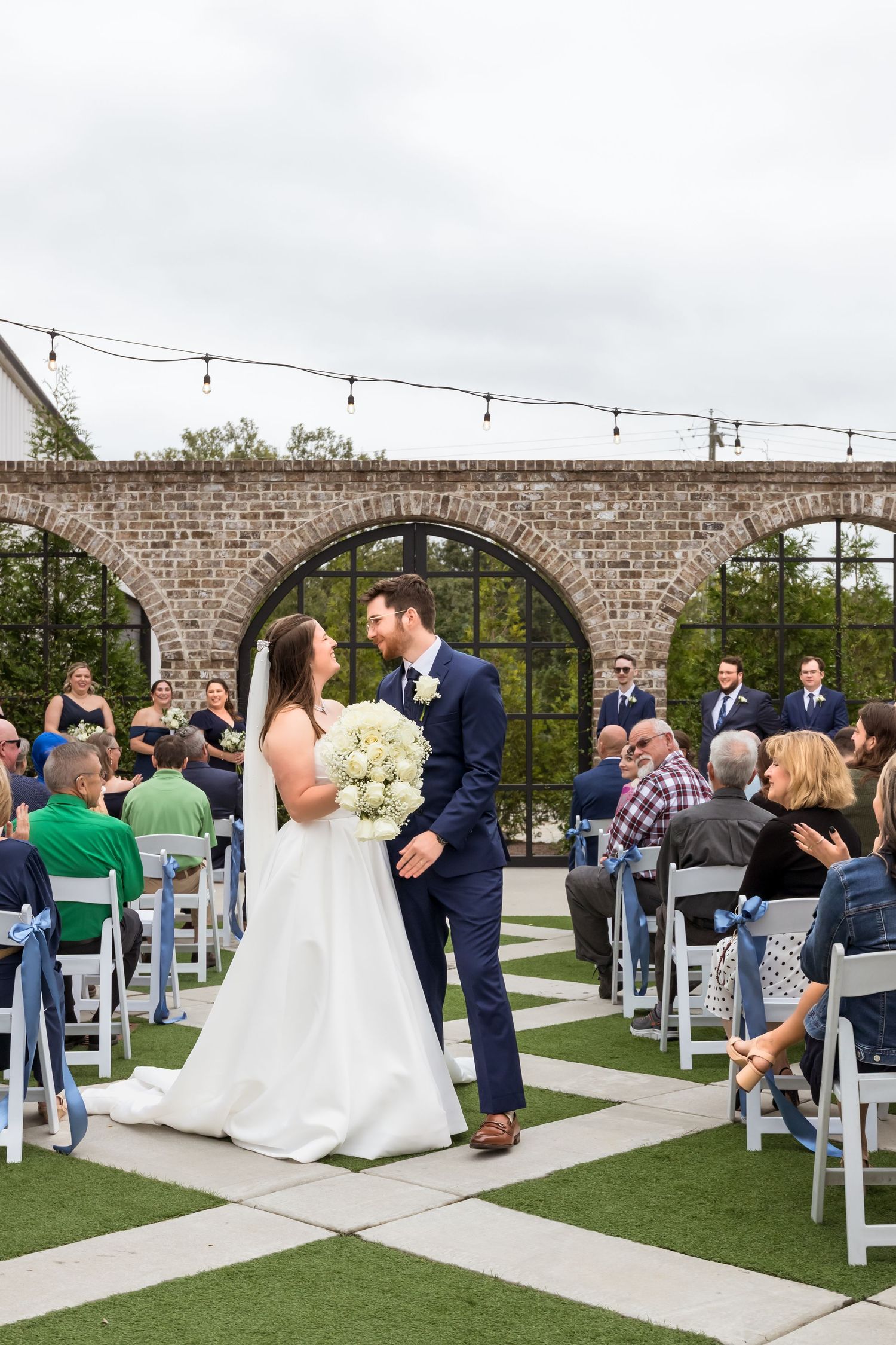 Outdoor wedding ceremony under a stone archway with white chairs and string lights.