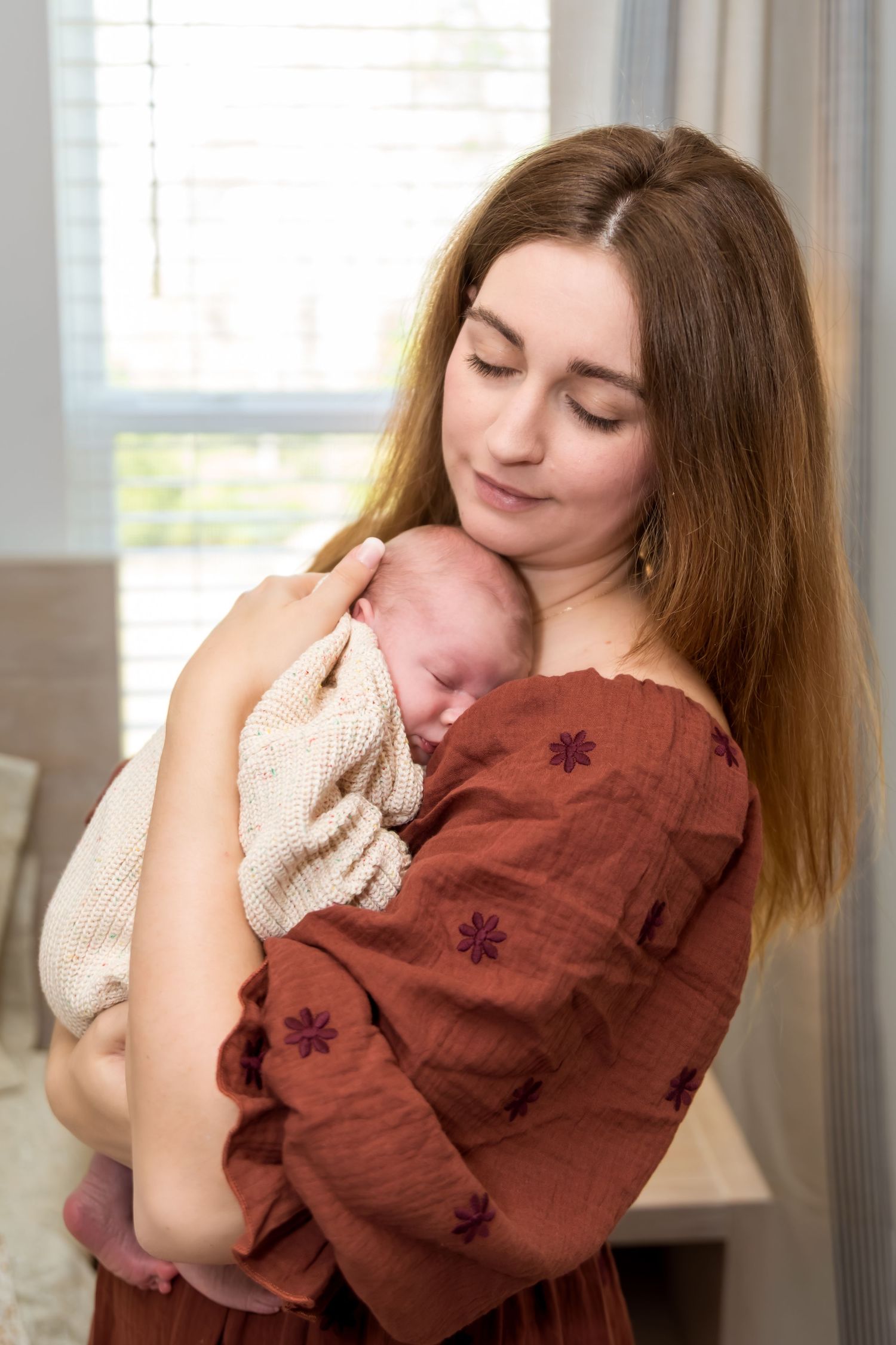 Young mother tenderly holds sleeping newborn baby wrapped in cream knit blanket, wearing rust floral dress.