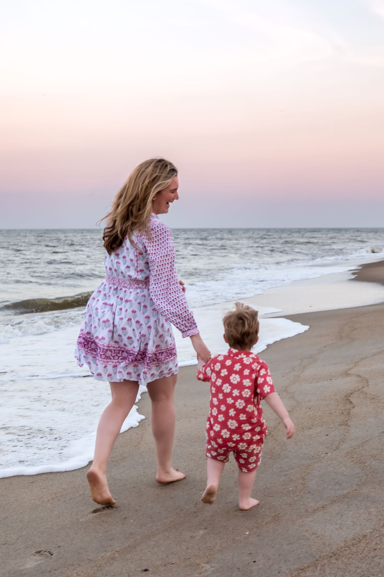 Mother and toddler walking hand in hand on a sandy beach at sunset, wearing matching floral outfits.