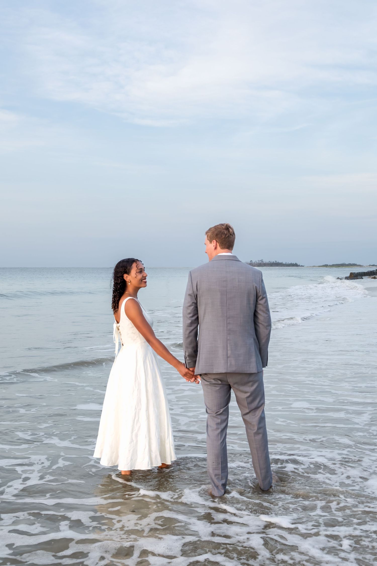Newlywed couple holding hands standing in ocean waves on beach, bride in white gown and groom in gray suit.