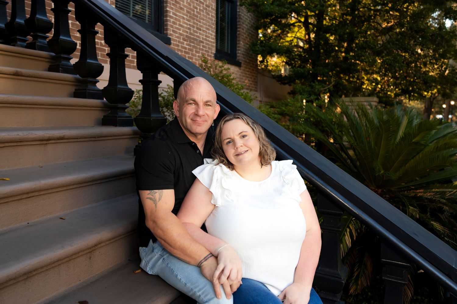 A couple sits together on outdoor brownstone steps during golden hour with palm plants nearby.