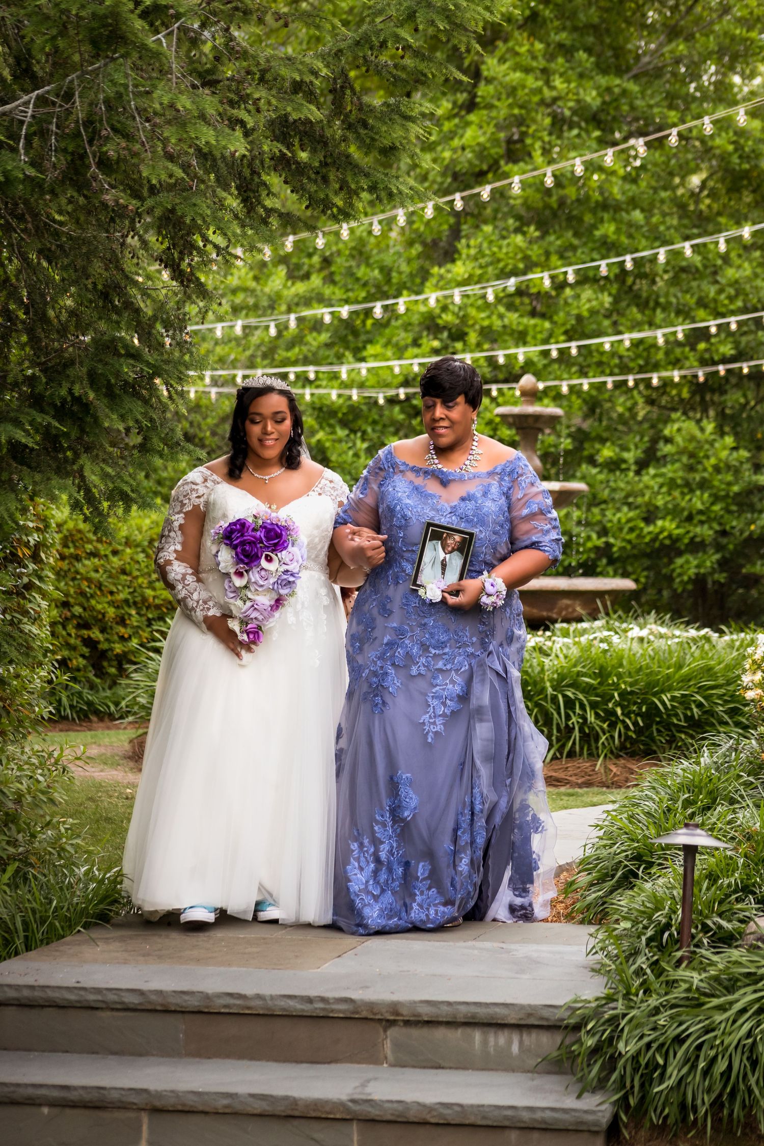 Two people in formal attire sit on stone steps decorated with string lights and greenery.