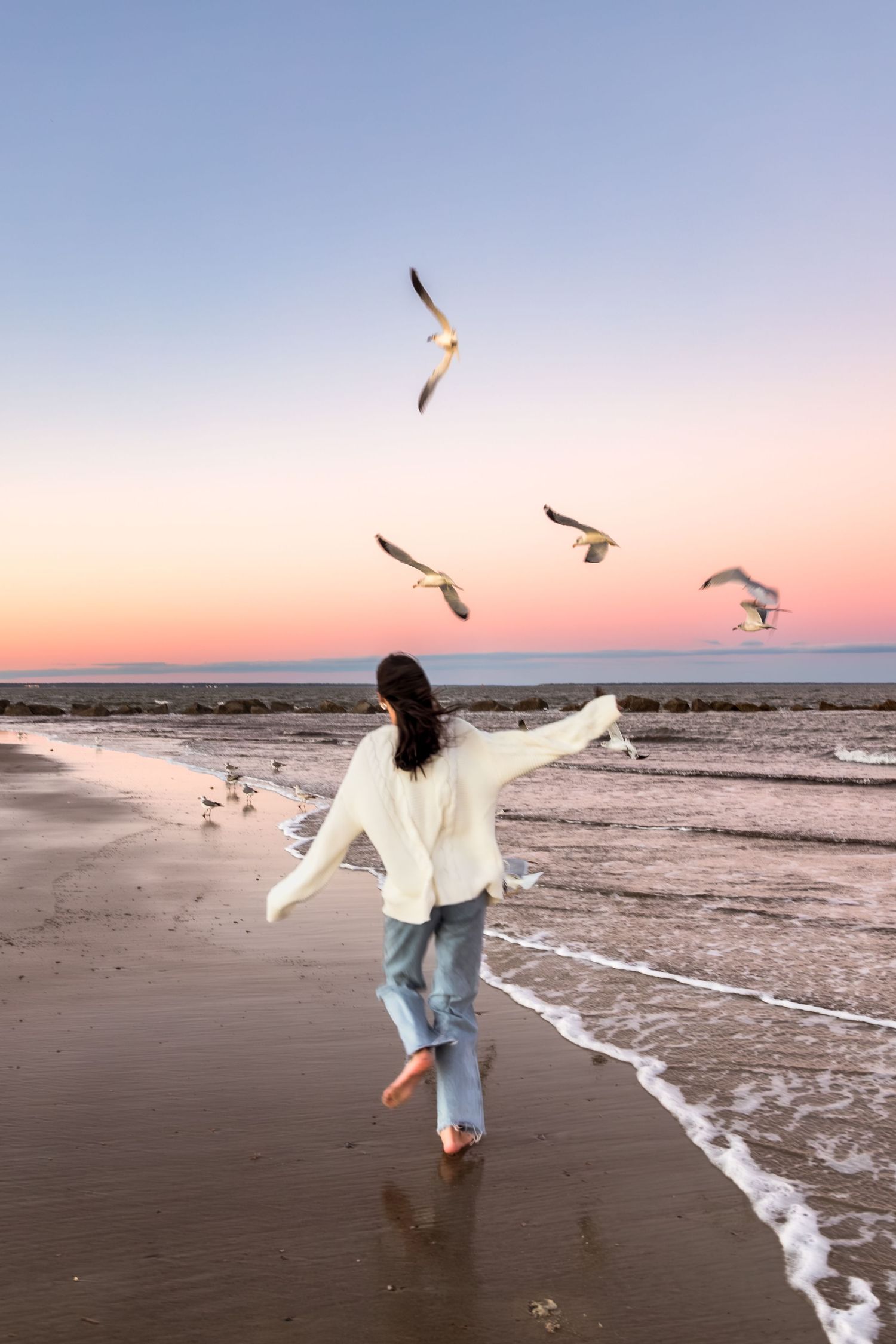 Person in white clothing running along beach at sunset while seagulls fly overhead.