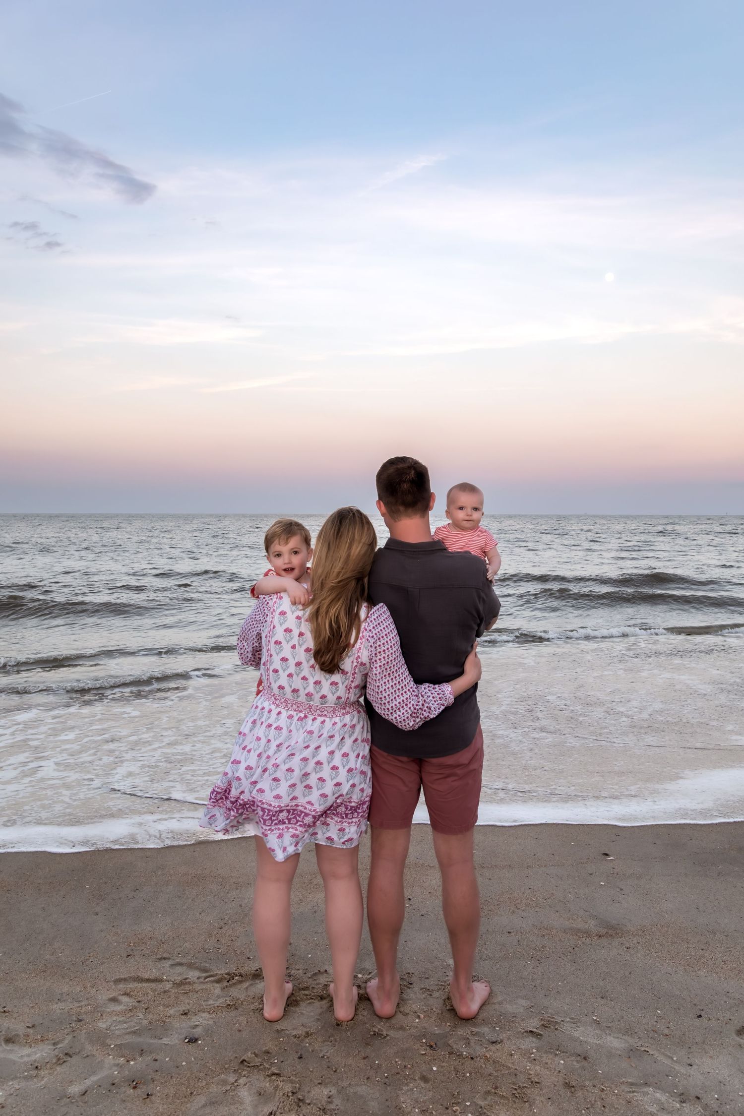 Family of four standing on beach at sunset, parents holding two young children while gazing at the ocean.