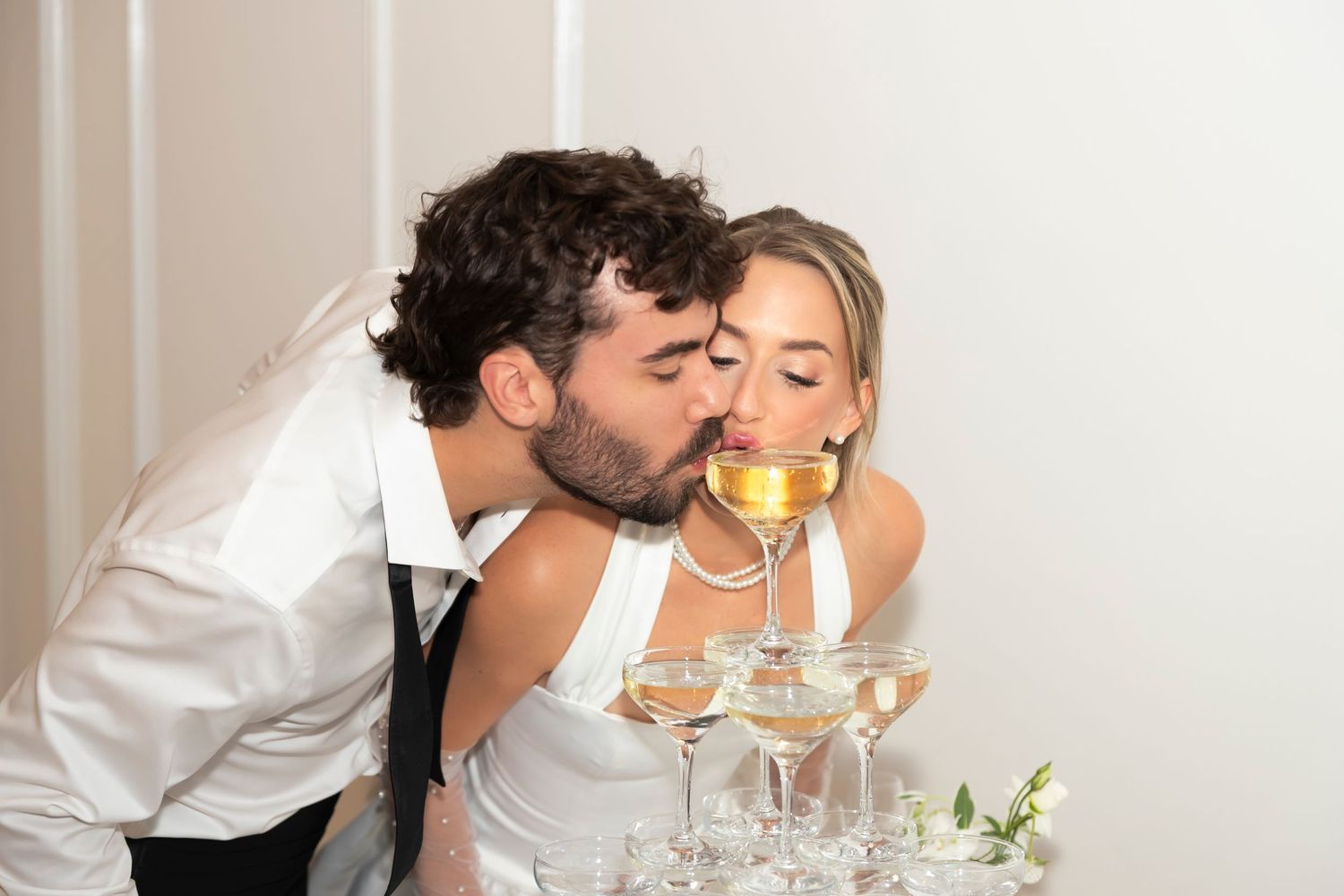 Couple sharing a romantic kiss while drinking champagne at a wedding reception with a champagne tower.