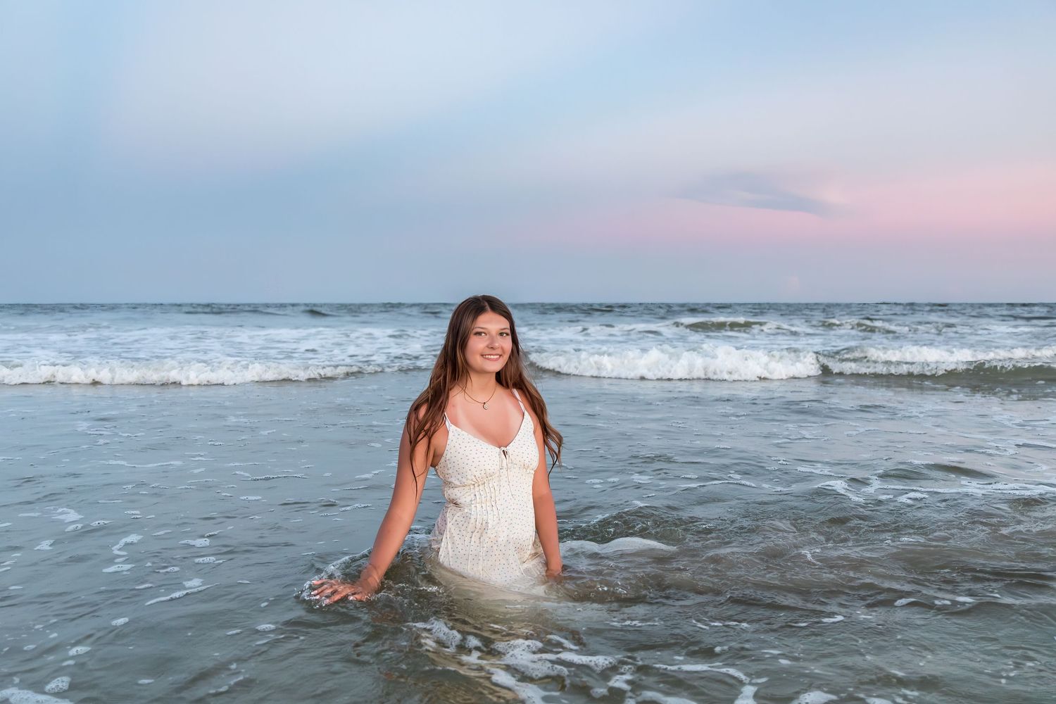 Young woman in white dress sitting in ocean waves at sunset on a beautiful beach.