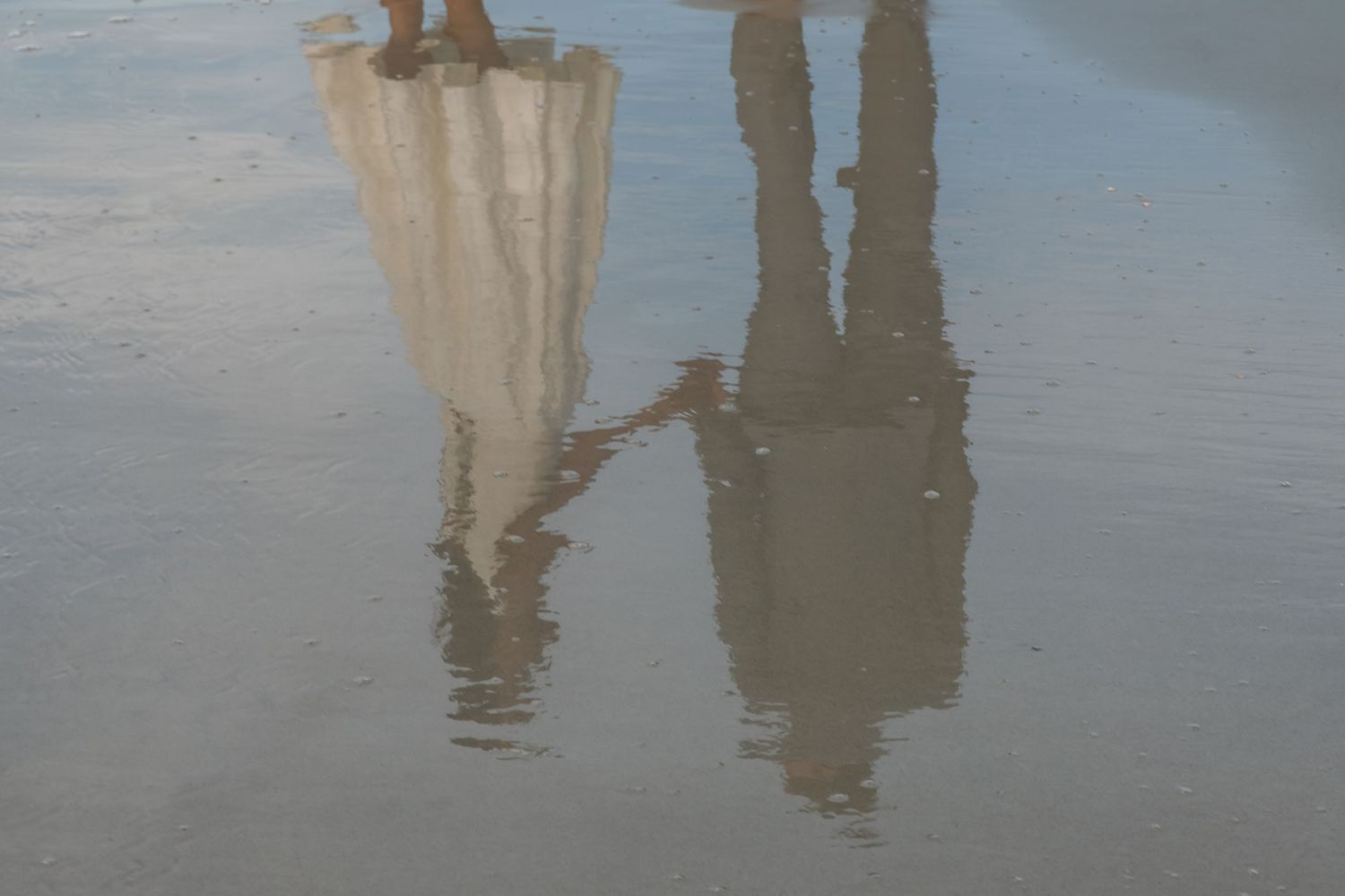 Reflection of bride in white dress and groom holding hands on wet sandy beach.