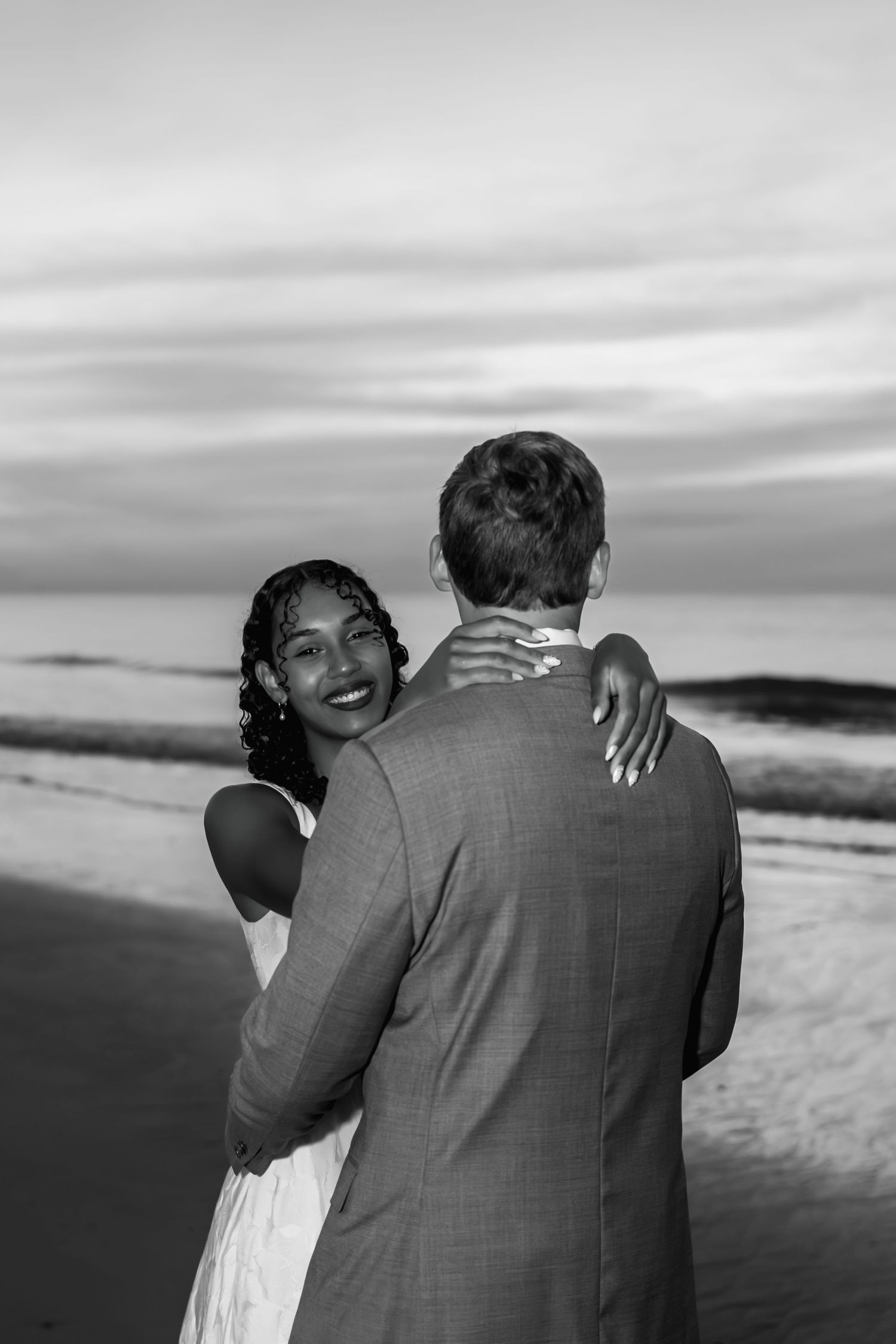 Black and white beach wedding photo of smiling bride embracing groom at sunset by the ocean.