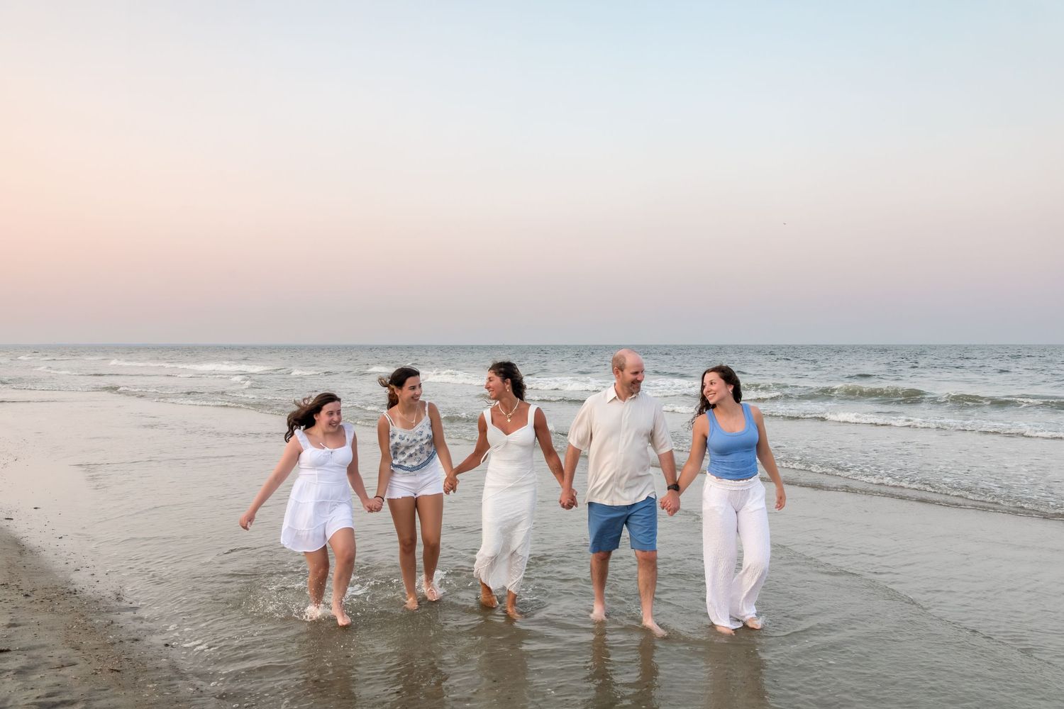 Family of five holding hands walking along a sandy beach at sunset in white and blue outfits.